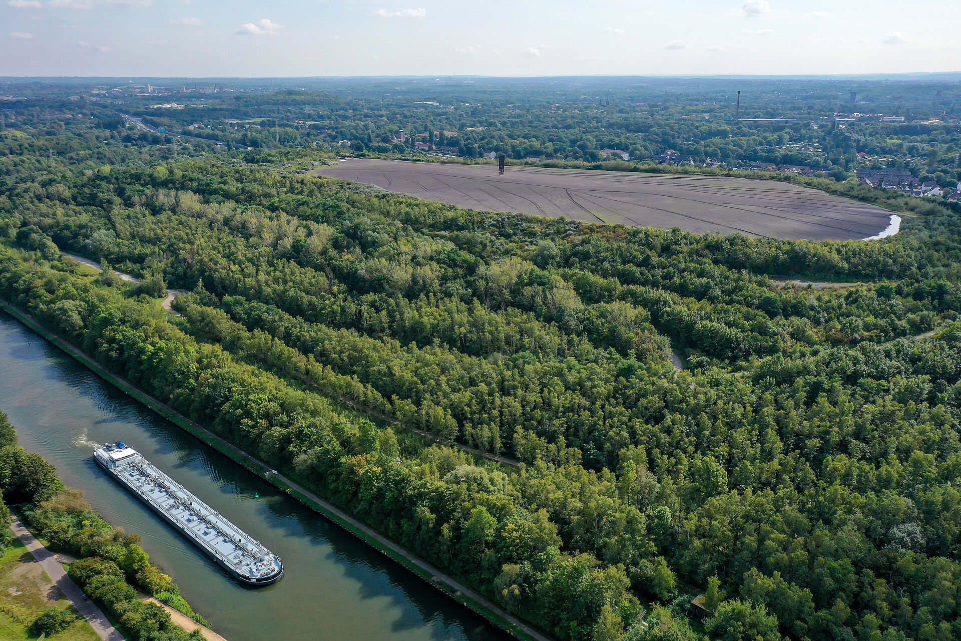 Im Vordergrund die Emscher, auf der ein Schiff fährt, dahinter ein Wald mit einer großen, freien Erd-/Baufläche in der Mitte.