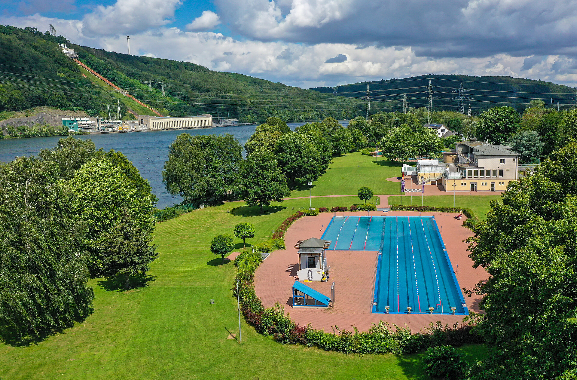 Vogelperspektive auf das Freibad umgeben von grünen Wiesen, direkt am Ufer des Hengsteysees. Im Hintergrund das Koepchenwerk auf der gegenüberliegenden Seite 
