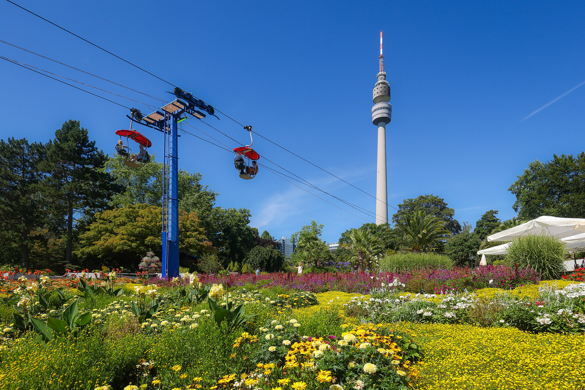 Ein Gondellift in einem Park mit bunten Blumen und im Hintergrund der Fernsehturm