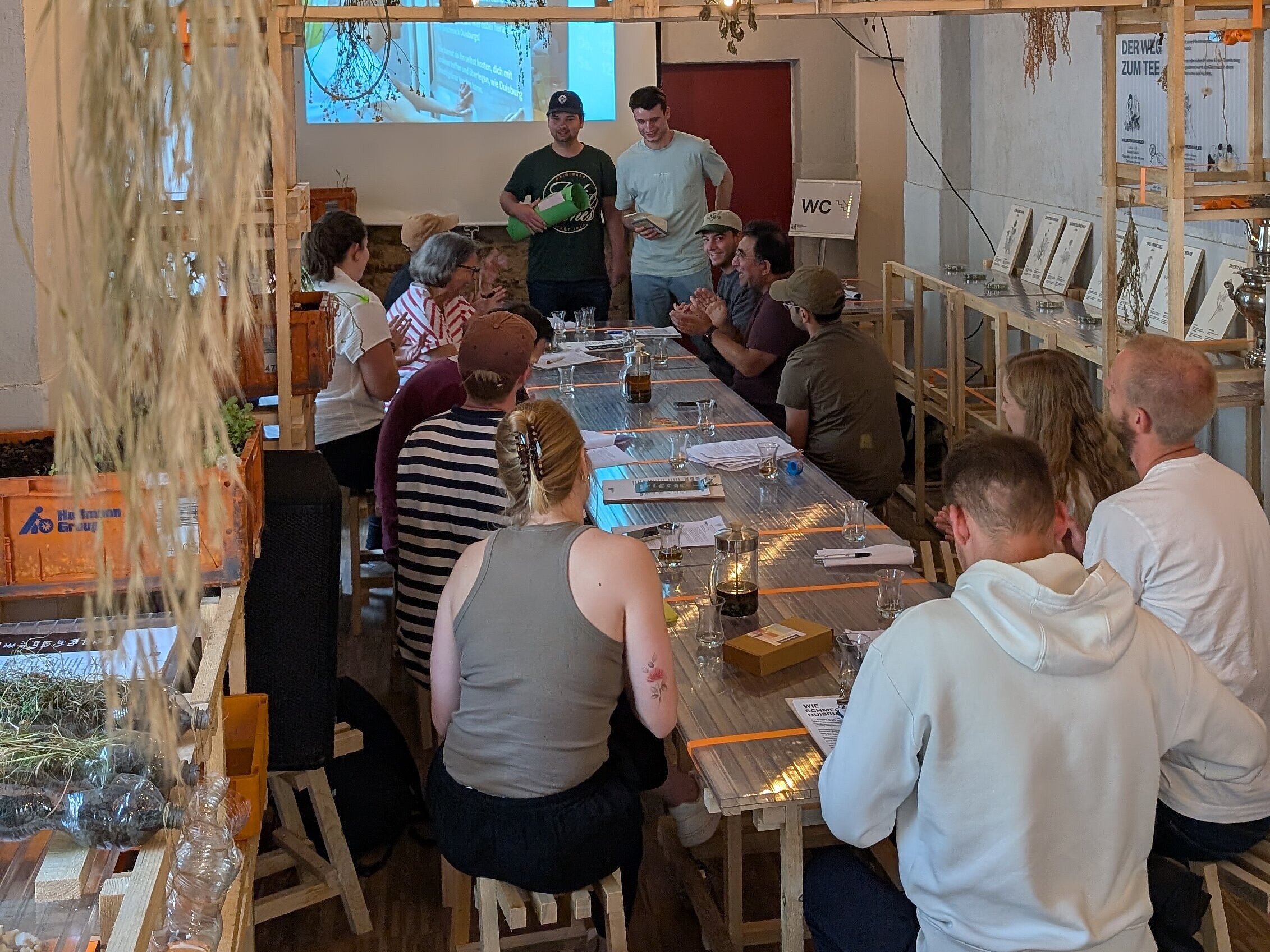 Innenansicht der Pop-Up-IGA Duisburg während eines Seminar der Ruhr-Universität-Bochum. Rückansicht von mehreren Studierenden, die an Tischen sitzen und auf eine Leinwand sehen, auf die etwas von einem Beamer projiziert wird.