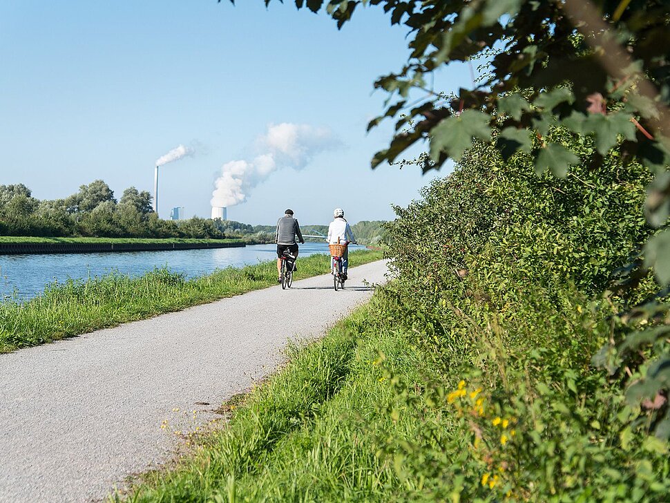 Das Bild zeigt zwei Fahrradfahrer, die an einem Fluss entlang radeln.