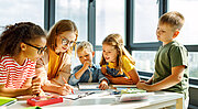 Eine Lehrkraft sitzt mit 4 Kindern an einem Gruppentisch und bearbeitet Aufgaben