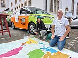 Mit dem Klimamobil Konsum hinterfragen Zwei Referent*innen des Klimamobils knien auf einer großen, begehbaren Weltkarte. Im Hintergrund das Kolping-Klimamobil.