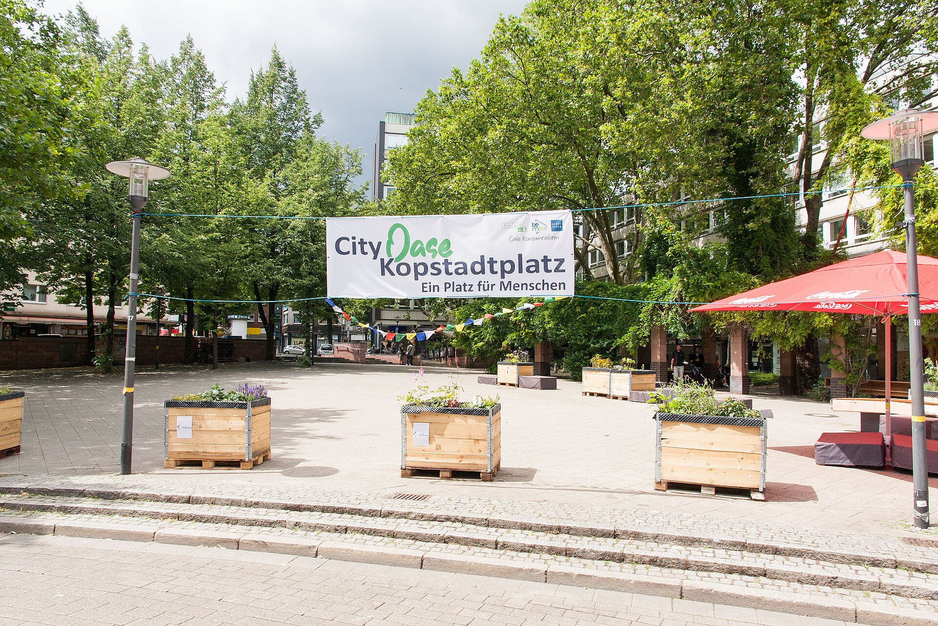 Foto eines gepflasterten Platzes in der Stadt. Im Vordergrund stehen Hochbeete aus Holz, im Hintergrund ist ein Banner mit der Aufschrift "City Oase" Kopstadtplatz gespannt.