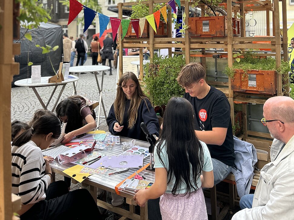 Außenbereich der Pop-Up-IGA Duisburg während des Straßenfests. Mehrere Jugendliche sitzen an einem Tisch und basteln. 