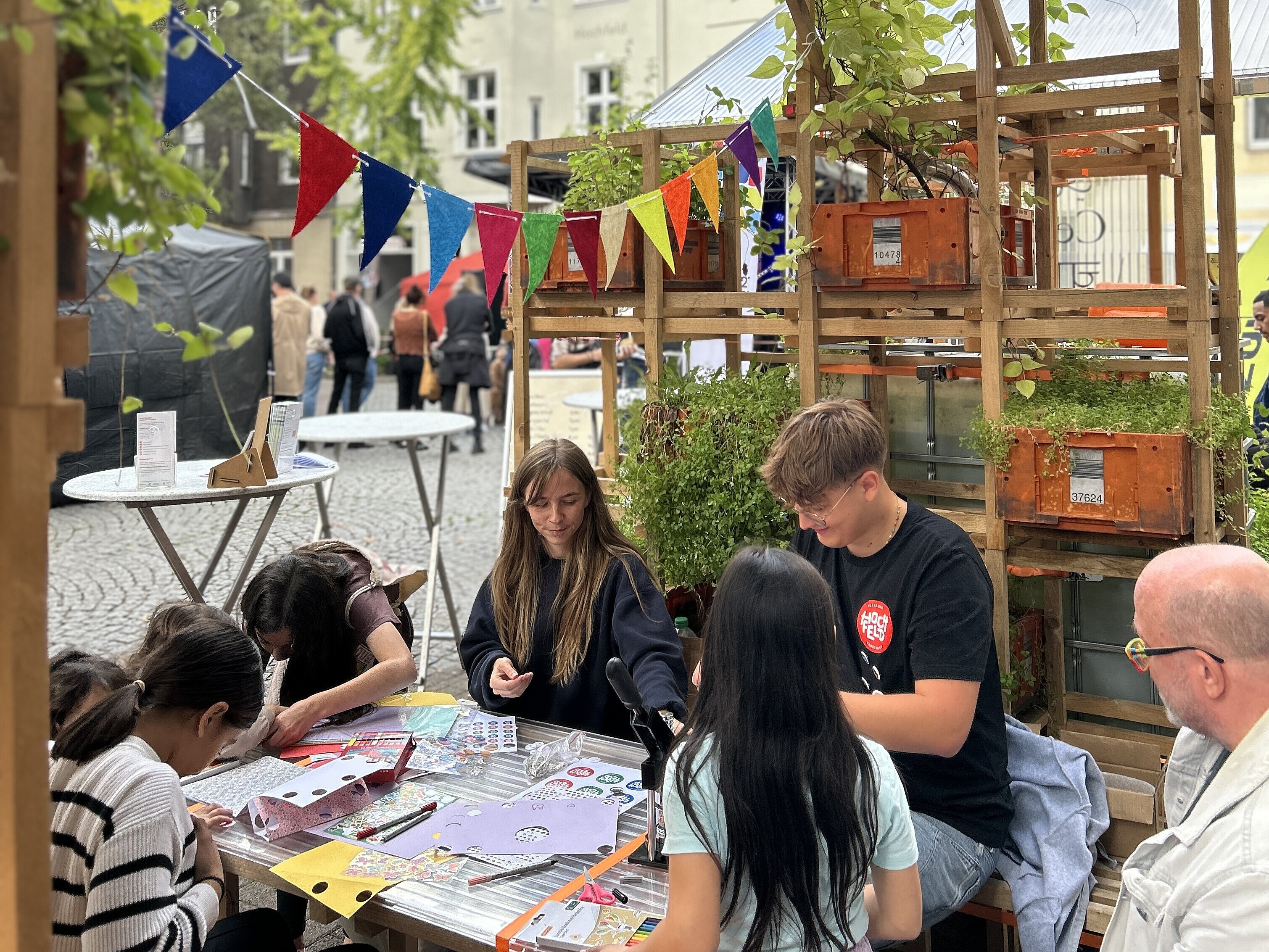 Außenbereich der Pop-Up-IGA Duisburg während des Straßenfests. Mehrere Jugendliche sitzen an einem Tisch und basteln. 