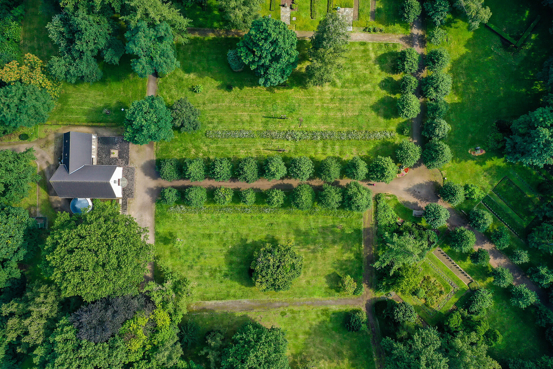 Drohnensicht auf ein Gebäude inmitten eines Waldes. Umgeben von grüner Wiese und einem Weg, der durch eine Baumallee herführt