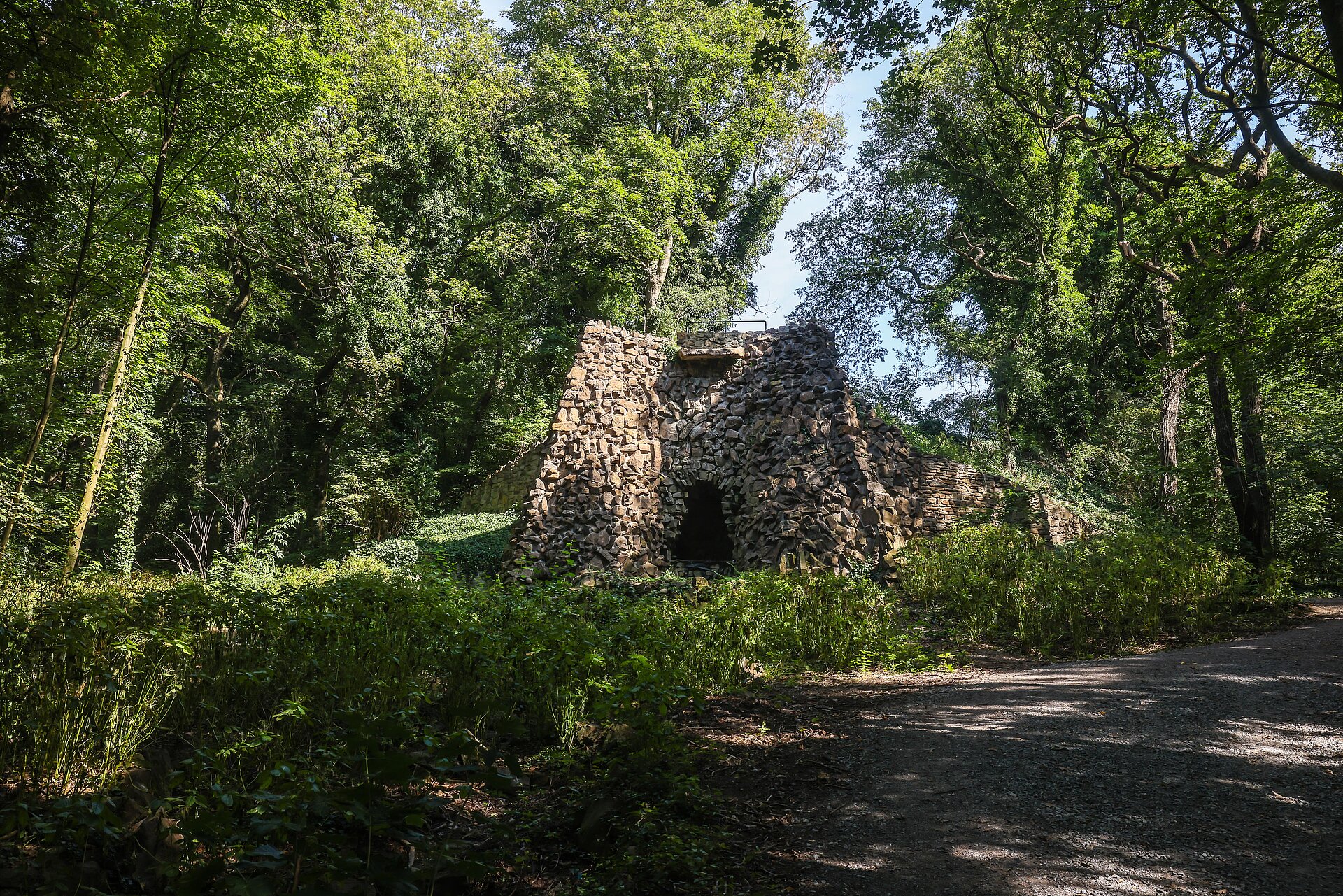 Mittig eine Steinmauer/Höhle mit Loch in der Mitte, an einem Waldweg, umgeben von Bäumen