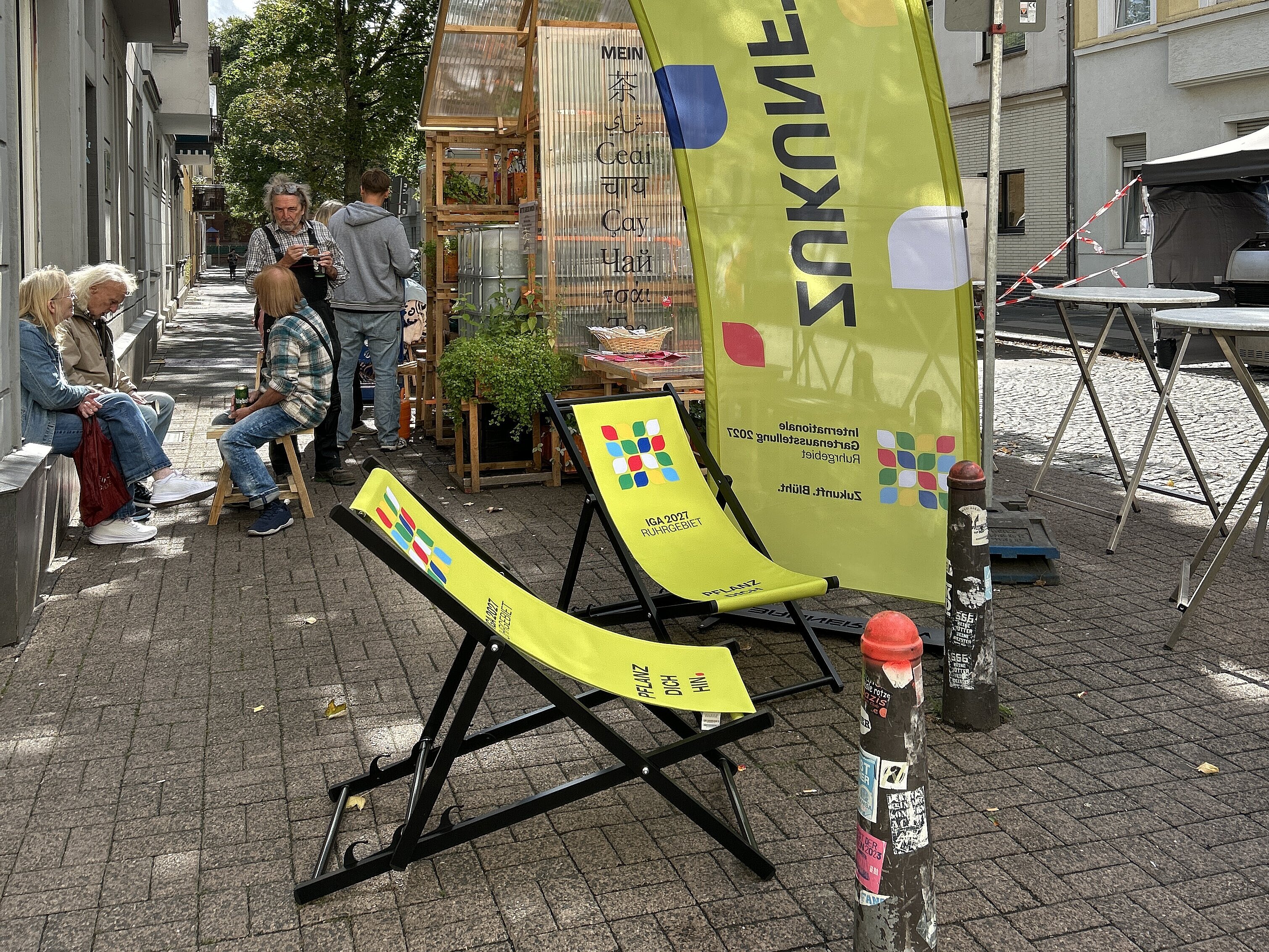 Außenbereich der Pop-Up-IGA Duisburg. Zwei grüne Liegestühle mit dem IGA-Logo überspannt von einem grünen Bannerbow mit IGA-Logo. Im Hintergrung sitzende Mesnchen, die sich unterhalten.