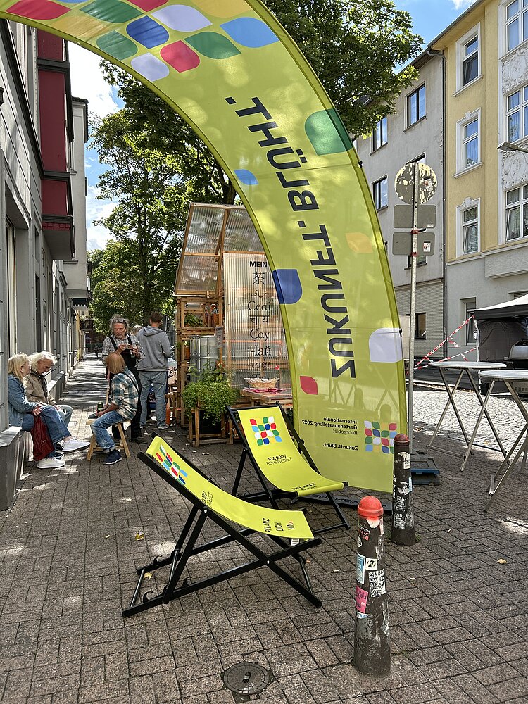 Außenbereich der Pop-Up-IGA Duisburg. Zwei grüne Liegestühle mit dem IGA-Logo überspannt von einem grünen Bannerbow mit IGA-Logo. Im Hintergrung sitzende Mesnchen, die sich unterhalten.