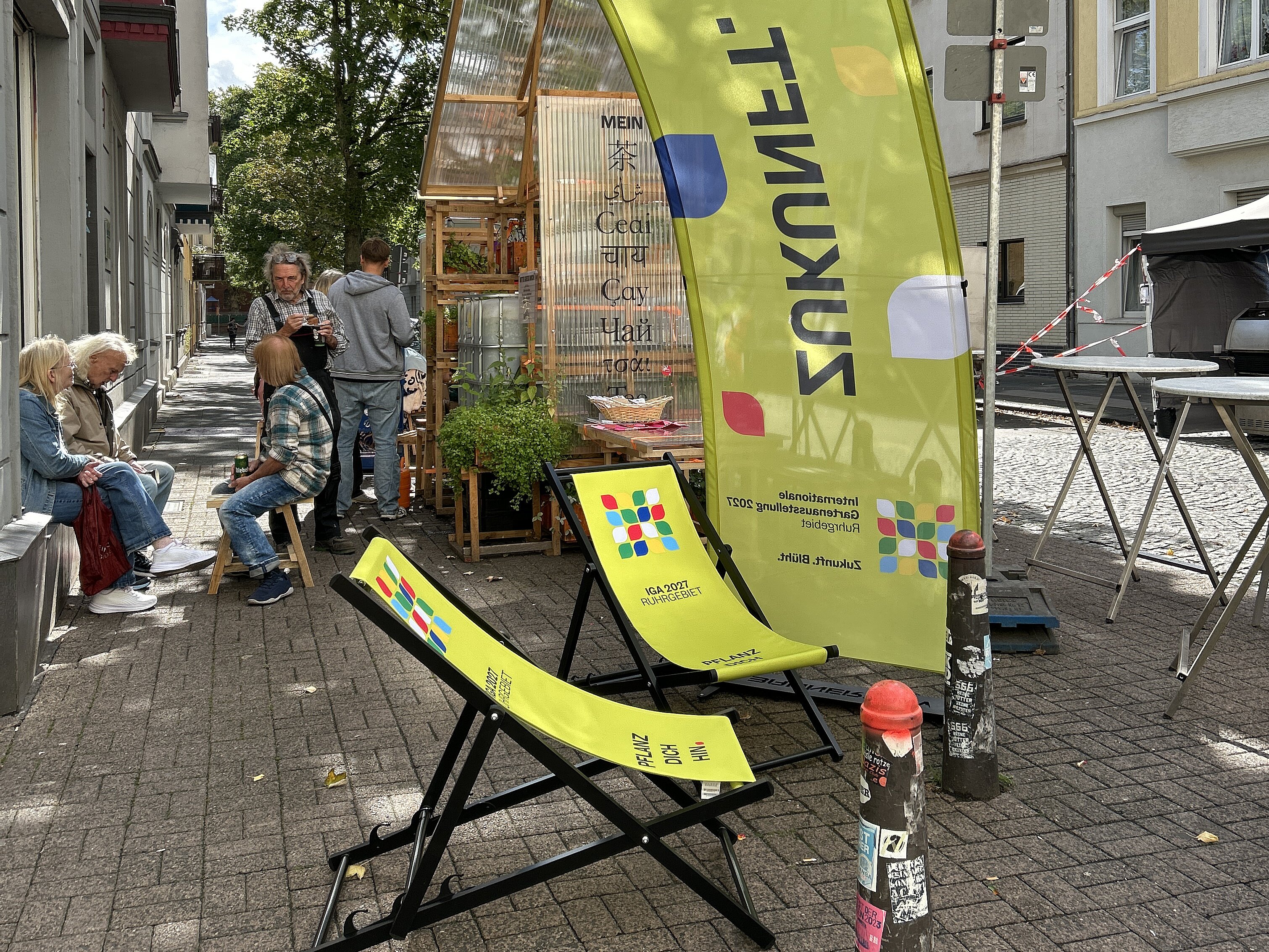 Außenbereich der Pop-Up-IGA Duisburg. Zwei grüne Liegestühle mit dem IGA-Logo überspannt von einem grünen Bannerbow mit IGA-Logo. Im Hintergrung sitzende Mesnchen, die sich unterhalten.
