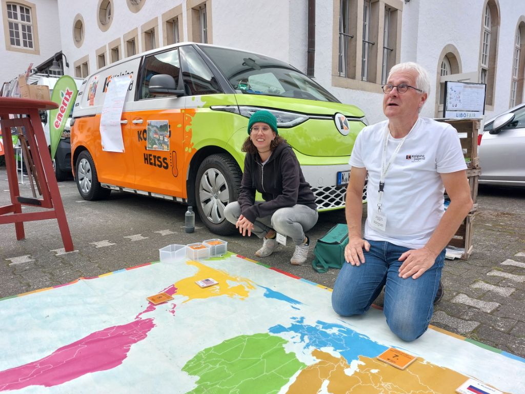KOLPING-Klimamobil_Paderborn_Kolping_2024 Zwei Referent*innen des Klimamobils knien auf einer großen, begehbaren Weltkarte. Im Hintergrund das Kolping-Klimamobil.