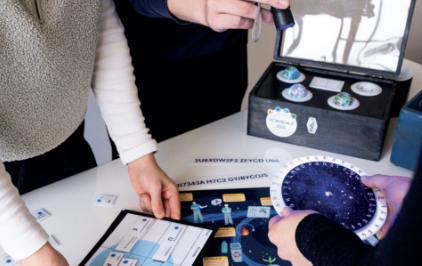 Zirkulär im Orbit_Wuppertal_Neue Effizienz_2025 Zu sehen sind die Hände von drei Personen, die verschiedene spielerische Elemente, beispielsweise eine drehbare Codierscheibe, ausprobieren. Im Hintergrund steht eine Holzkiste auf dem Tisch, in welcher sich vier bunter Kugeln befinden.