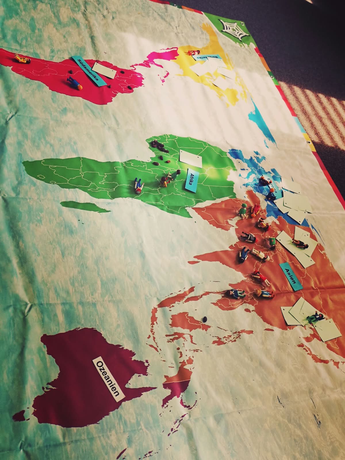 Weltfairteilung_Marchen_Chrost_2026 Fotografie einer Weltkarte auf einem Tuch. Die Kontinente sind farbig markiert und mit kleinen Spielfiguren, Karten und Beschriftungen versehen. Sichtbar sind die Bezeichnungen „Stammamerika“, „Afrika“, „Asien“ und „Ozeanien“. Die Darstellung wirkt wie eine Gruppen- oder Lernaktivität zur Verteilung der Weltbevölkerung bzw. von Ressourcen auf die Kontinente.
