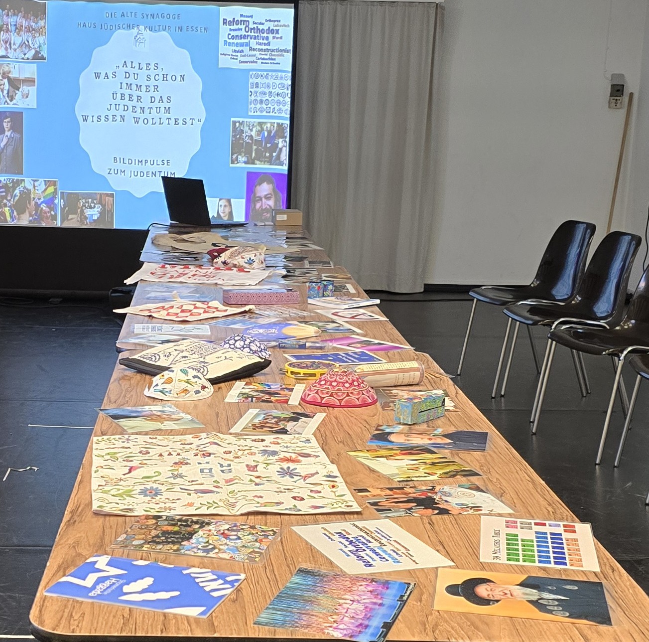 Workshop Alles was Du schon immer über Jüd*innen und Judentum wissen wolltest Das Foto zeigt einen großen Tisch mit zahlreichen Judaika-Stücke sowie Bildern, die das jüdische Leben an verschiedenen Orten darstellen.