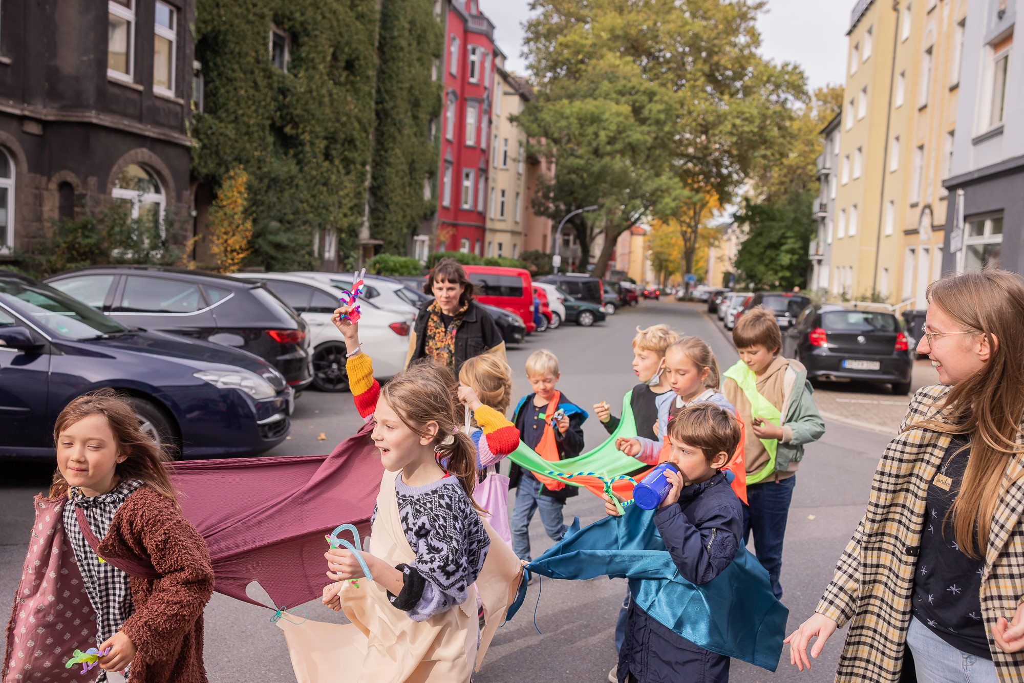 aufameisenwegen_dortmund_baege_2023 Eine Gruppe von Kindern bewegt sich gemeinsam als „Ameisenkette“ durch eine Straße. Die Kinder tragen farbige Tücher und bewegen sich miteinander verbunden zwischen parkenden Autos; eine erwachsene Begleitperson geht daneben. Ein Projekt vom Frappanz e. V