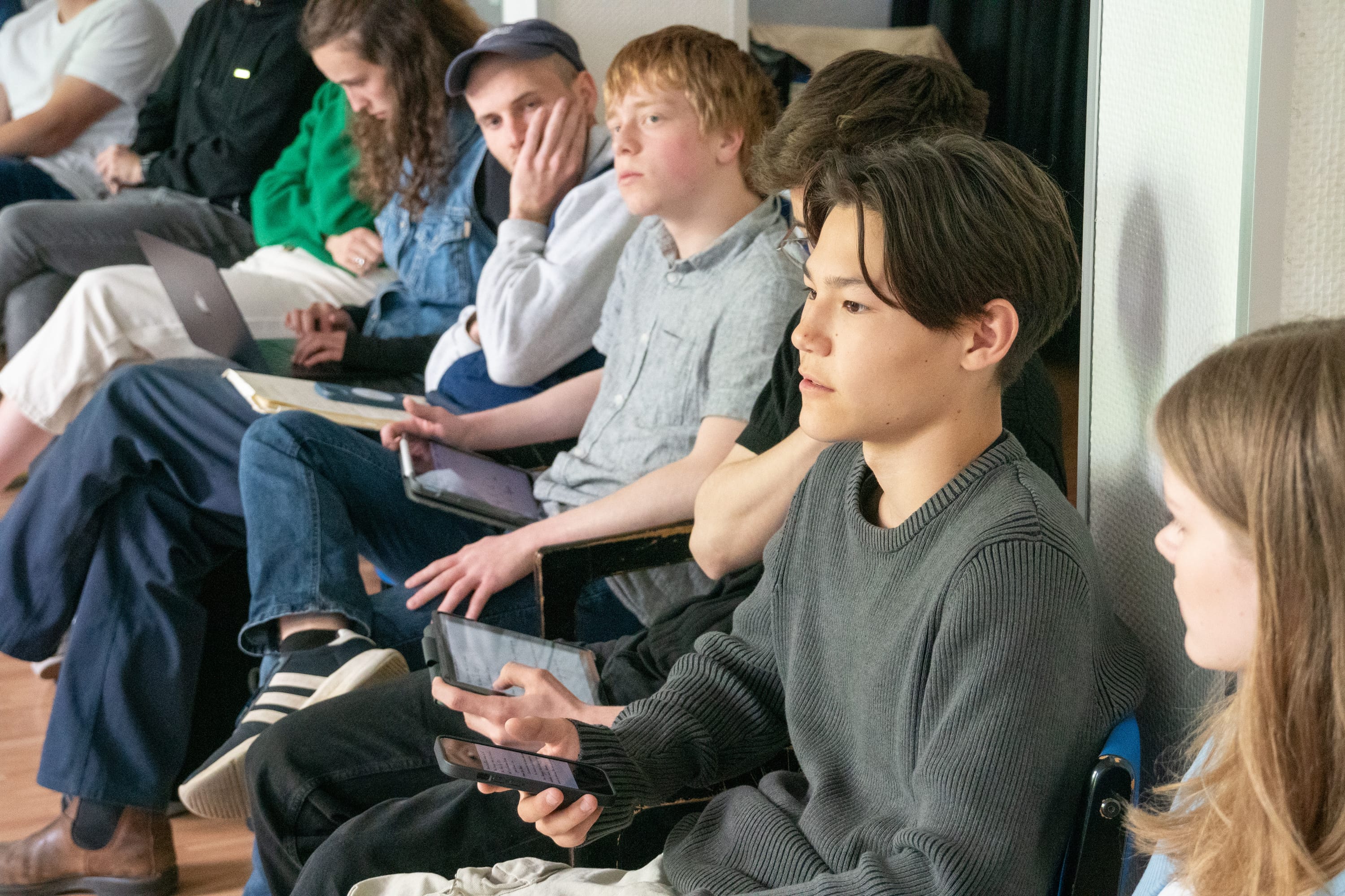Diskussion2_youmocracy e.V._2024 Eine Person argumentiert mit einem Smartphone in der Hand. Andere Personen hören aufmerksam zu und verfolgen die Ausführungen.
