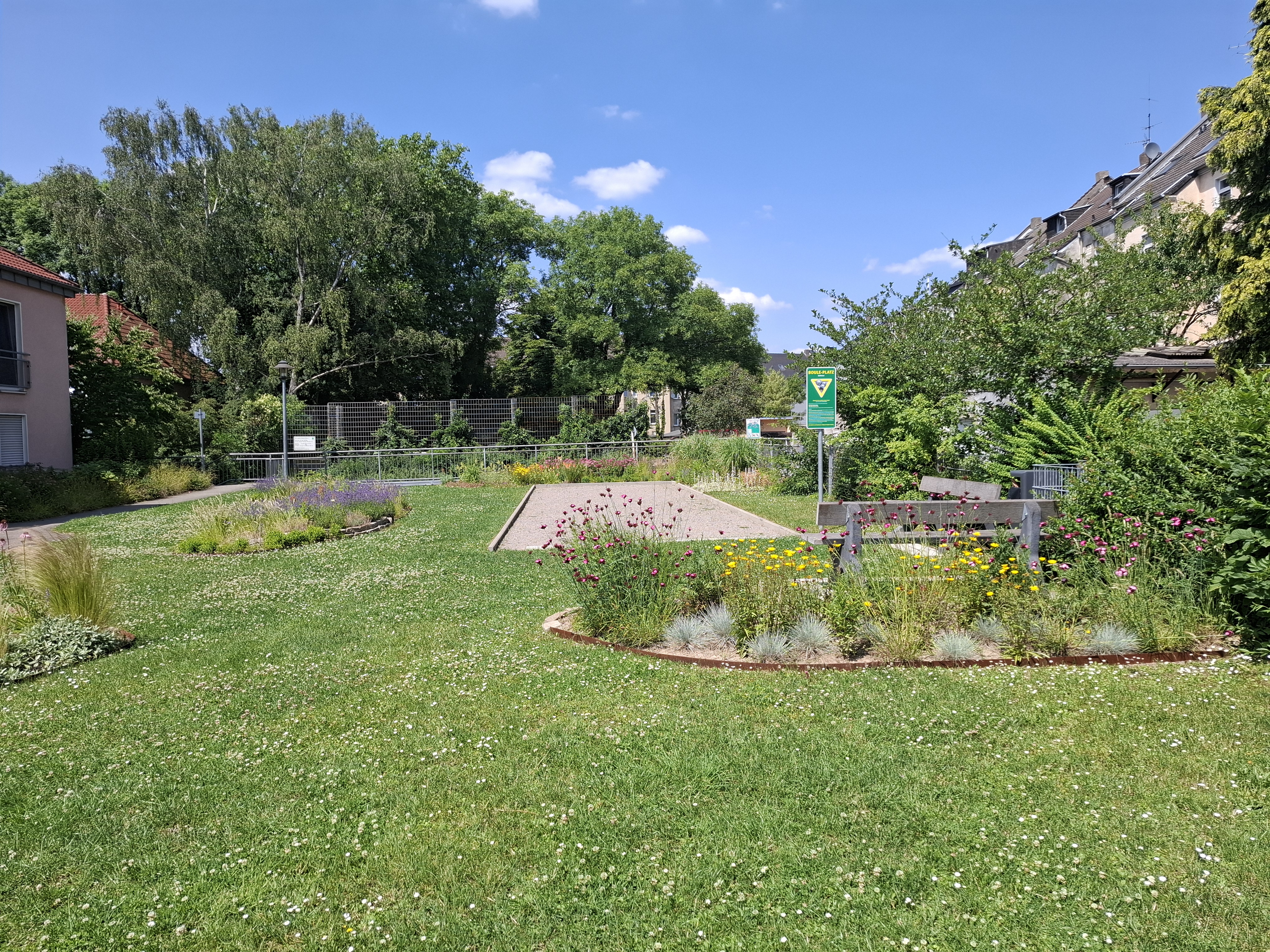 Für Anwohner*innen nutzbare Grünfläche inmitten von Wohnbebauung mit Blumenstreifen und Bouleplatz