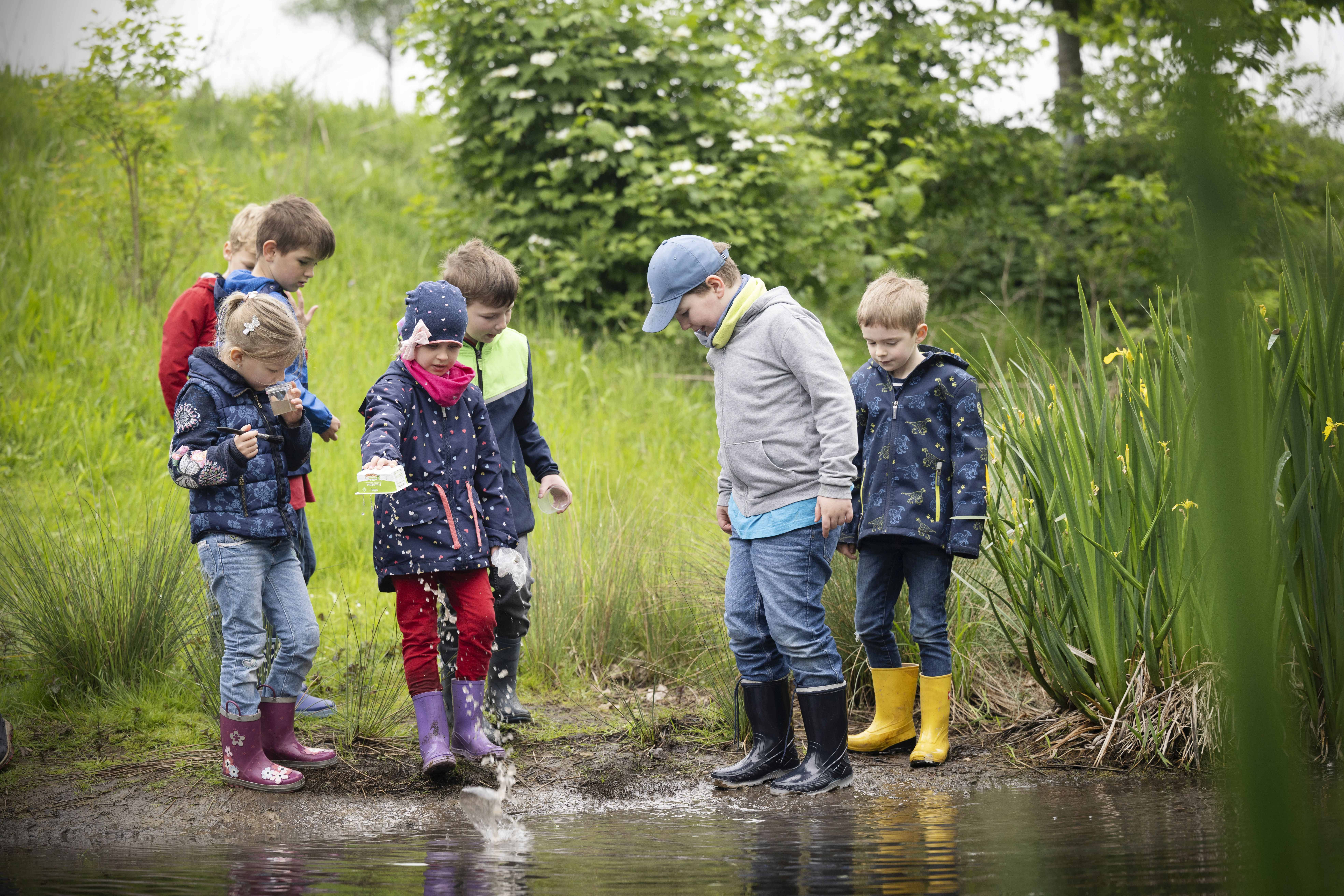 Zu sehen sind Kinder, die mit Regenklamotten und Gummistiefeln durch eine Pfütze im Grünen waten und sich diese anschauen