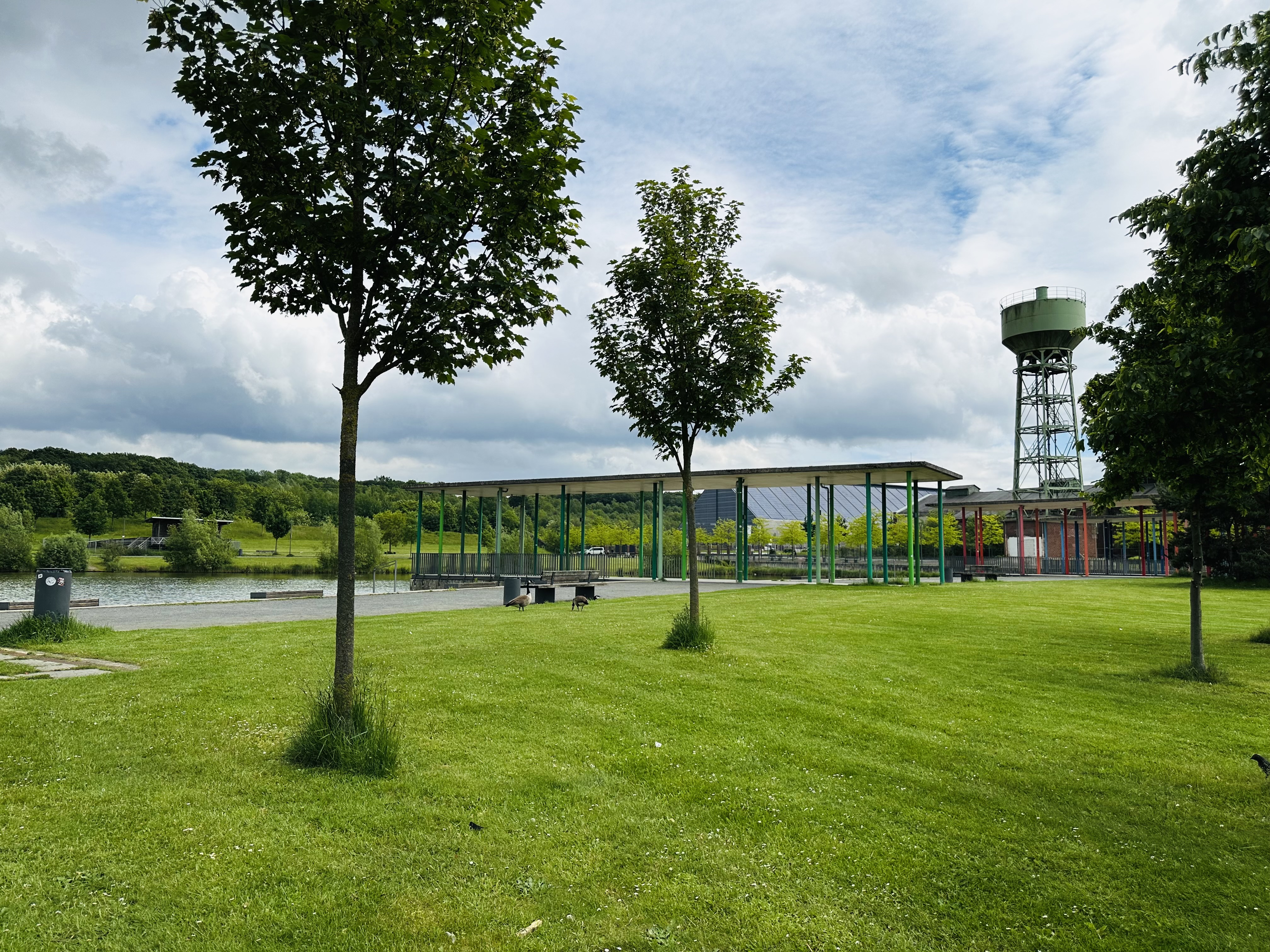 Im Vordergrund ist eine saftige grüne Rasenfläche mit jungen Bäumen zu sehen. Im Hintergrund befindet sich ein Teich, ein moderner Pavillon an dessen Ufer und ein Wasserturm.