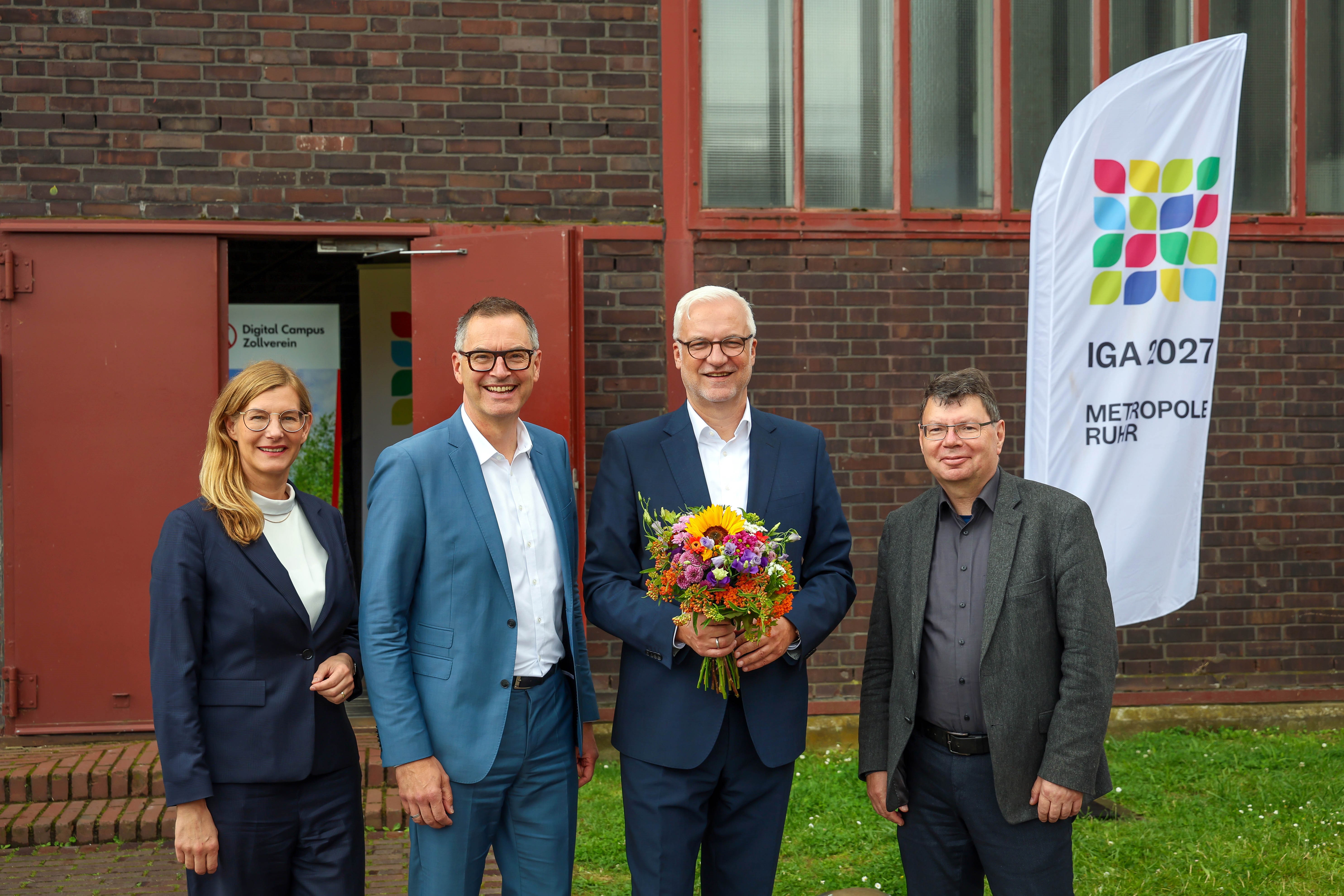Vier Personen stehen vor dem Gebäude des Digital Campus auf Zollverein, im Hintergrund eine Beachflag mit IGA-Logo und Aufschrift IGA 2027 Metropole Ruhr