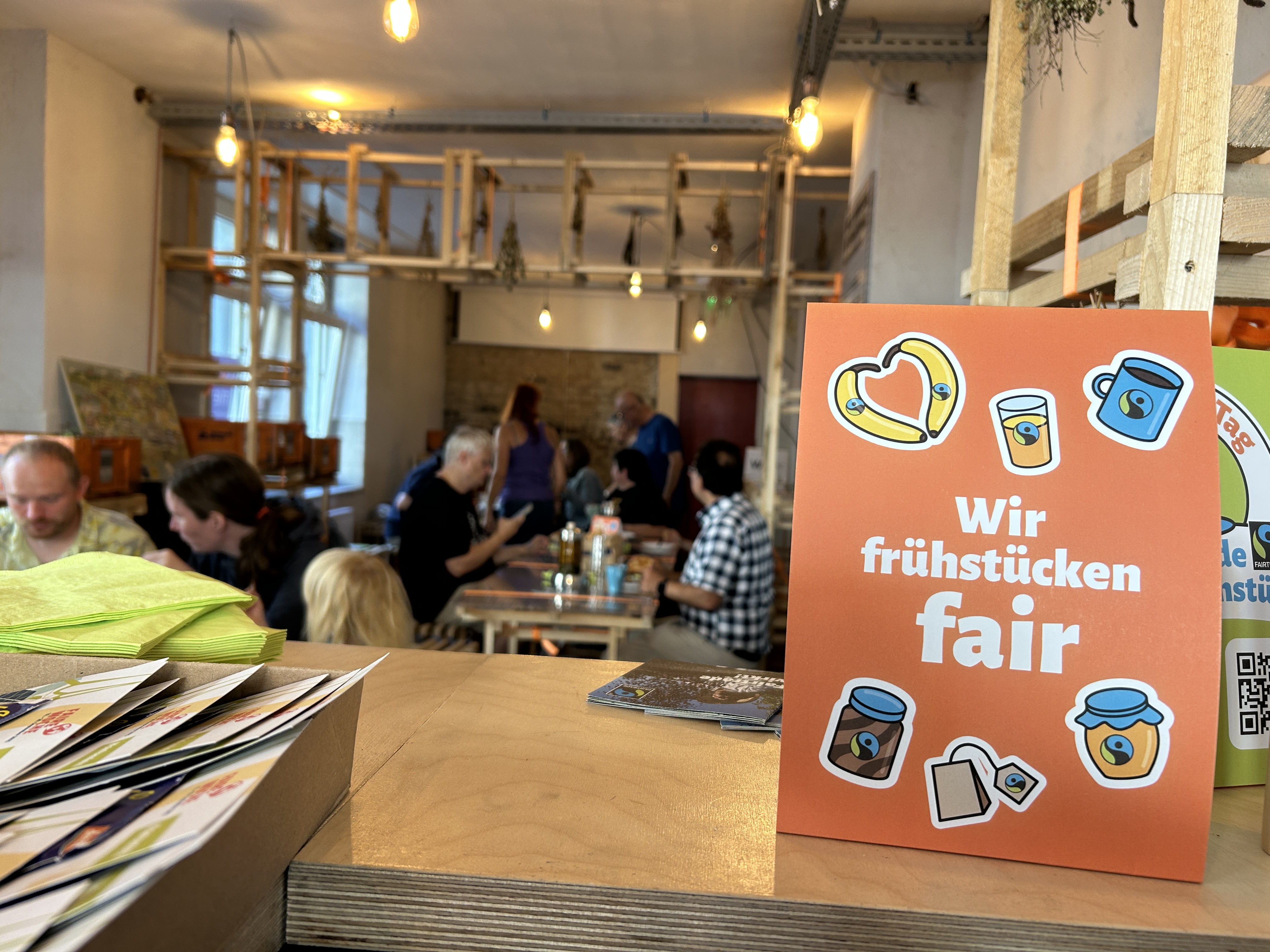 Innenraum der Pop-Up-IGA Duisburg. Im Vordergrund ein orangener Aufsteller auf dem Tresen mit der Aufschrift "Wir frühstücken fair". Im Hintergrund Menschen sitzend an Tischen beim essen.
