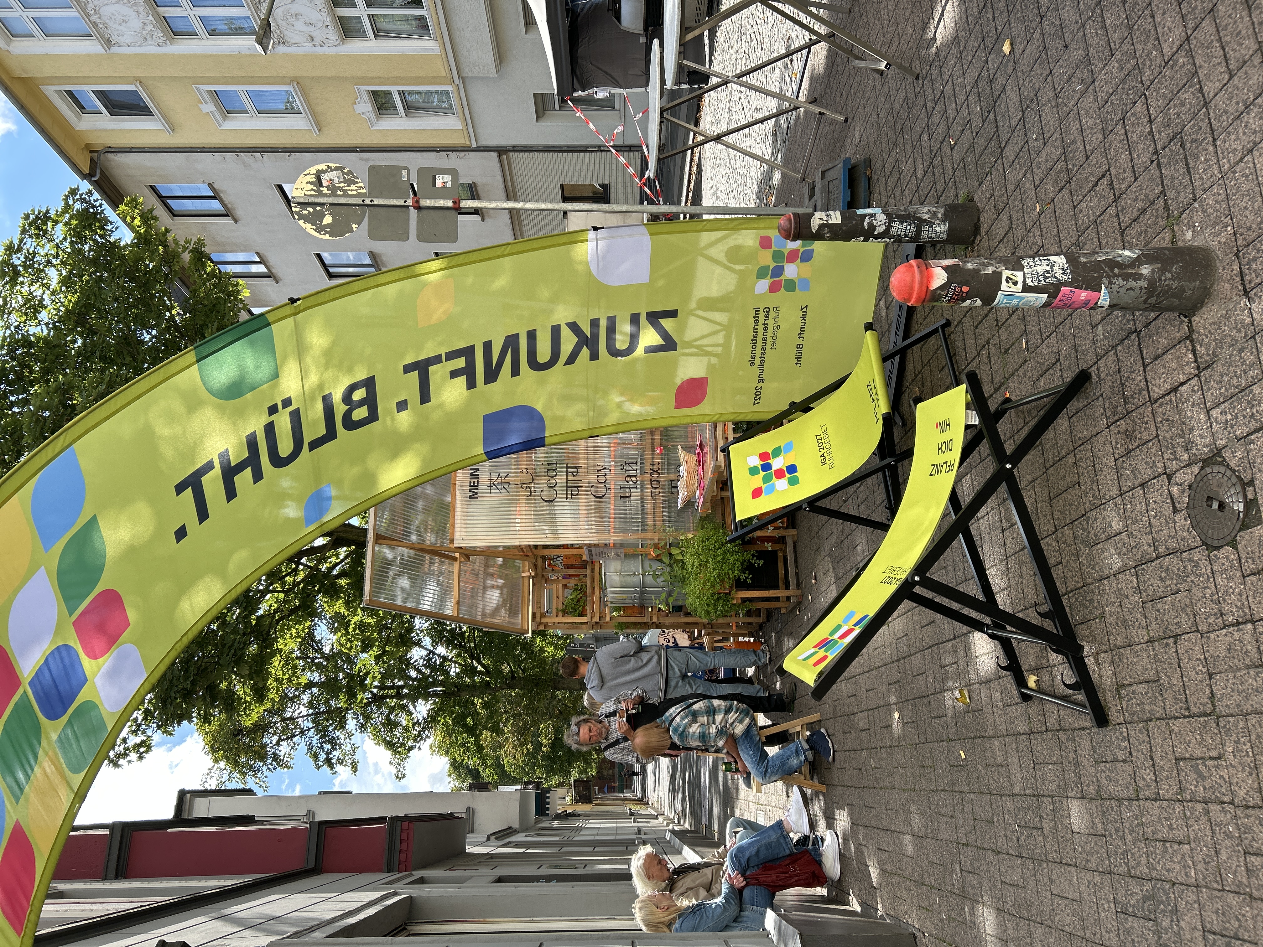 Außenbereich der Pop-Up-IGA Duisburg. Zwei grüne Liegestühle mit dem IGA-Logo überspannt von einem grünen Bannerbow mit IGA-Logo. Im Hintergrung sitzende Mesnchen, die sich unterhalten.