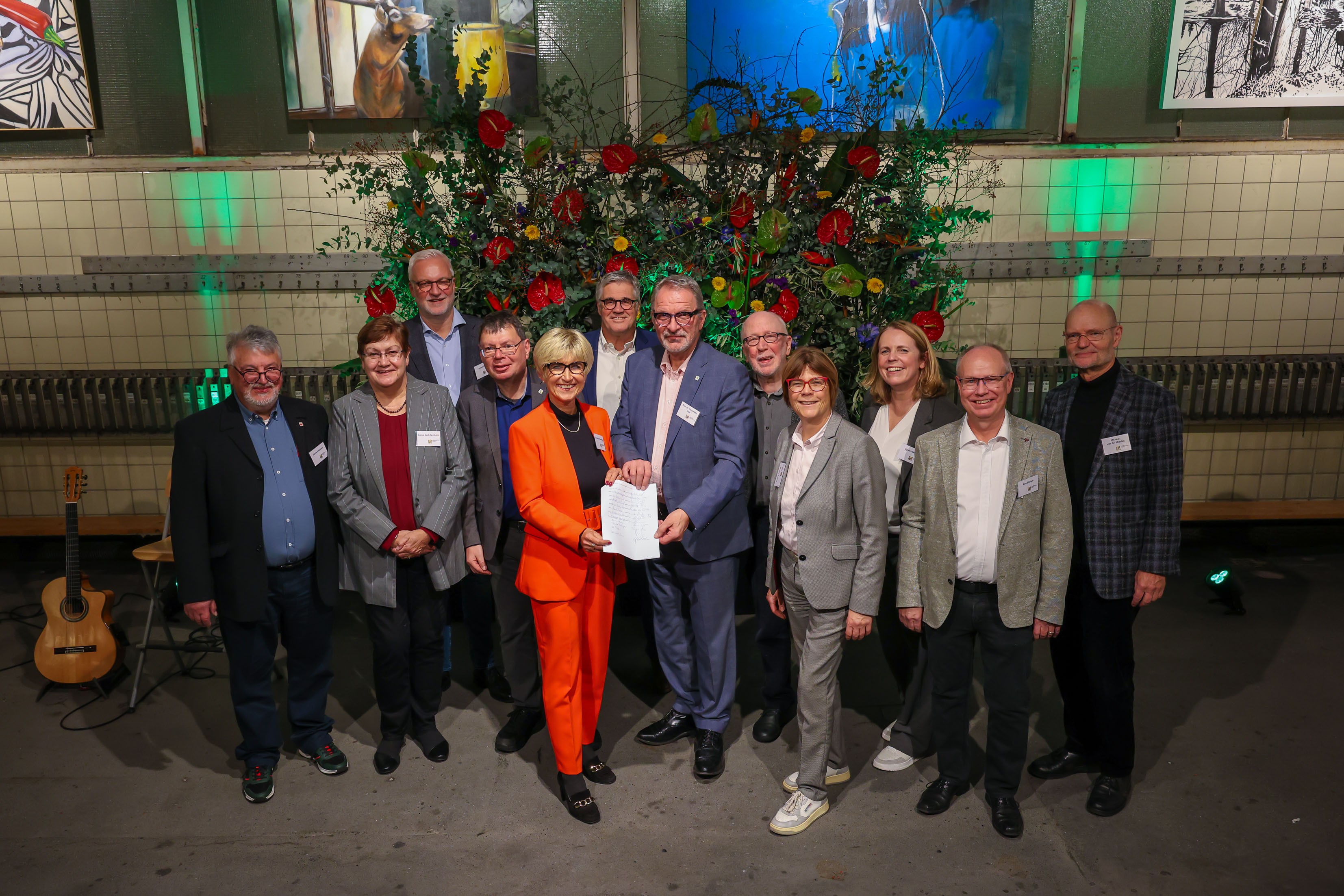 Vier Frauen und acht Männer stehen als Gruppe vor einer großen Blumendekoration und präsentieren ein Vertragspapier