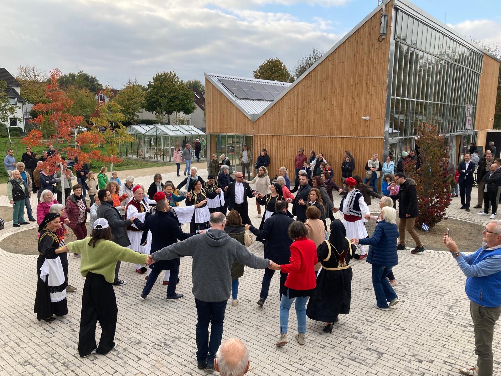 Eine Gruppe Menschen bei einem griechischen Tanz vor einem modernen Gebäude aus Holz und Glas in einem Park