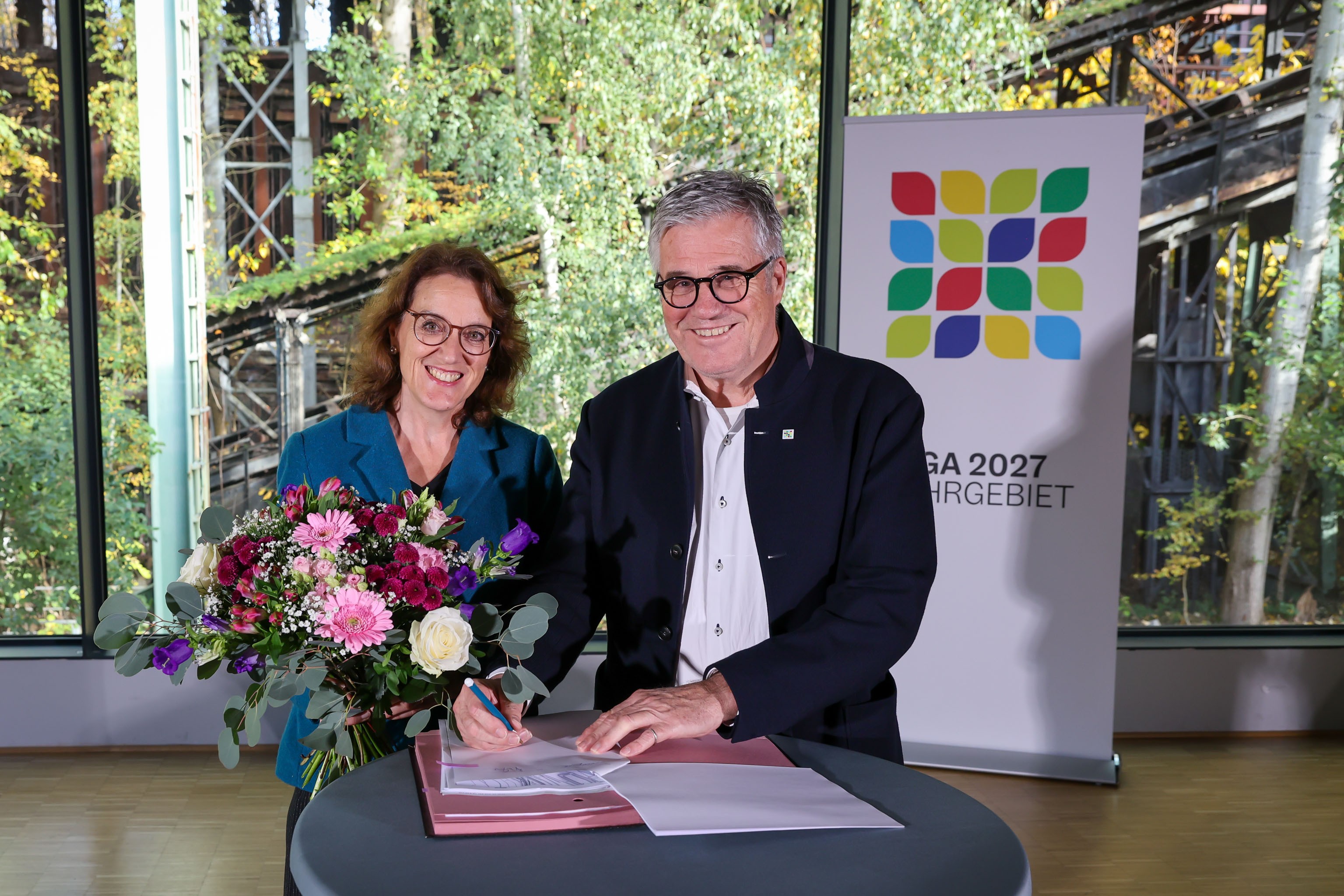 Eine Frau und ein Mann an einem Stehtisch einen Vertrag unterzeichnend vor einem großen Fenster, dahinter bewachsene Stahlkonstruktion