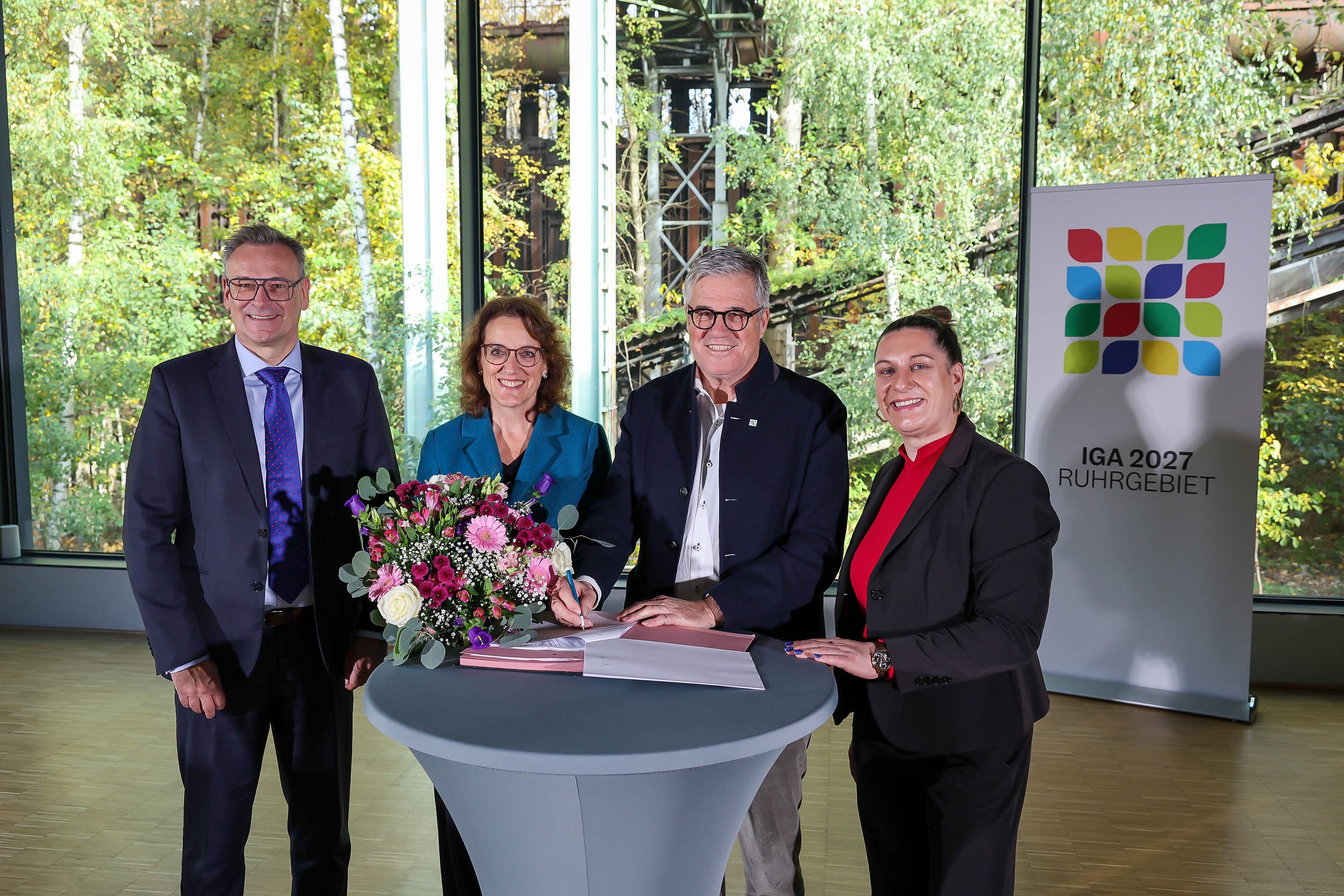 Zwei Männer und zwei Frauen an Stehtischen vor einem großen Fenster, dahinter bewachsene Stahlkonstruktion