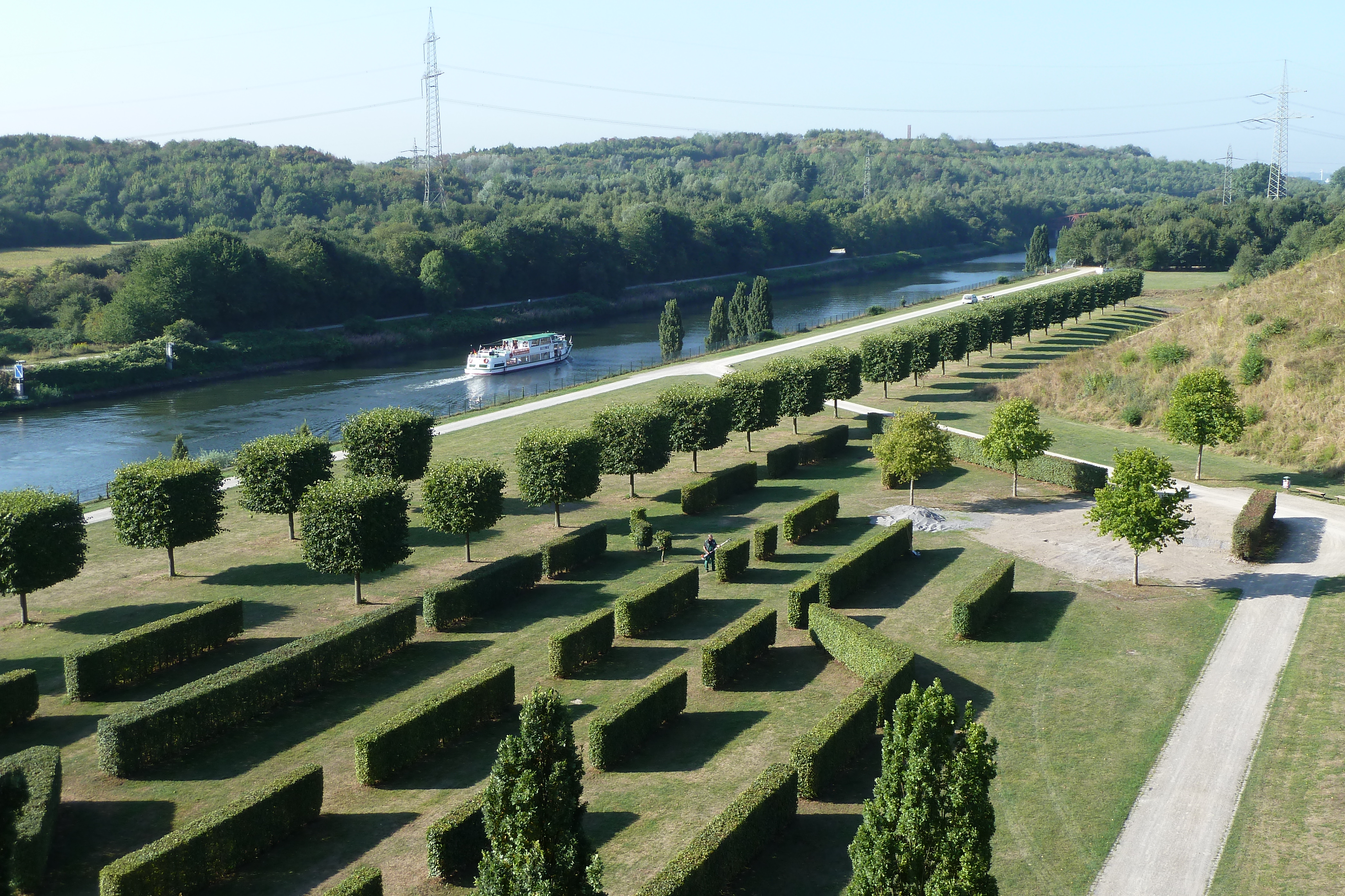 Das Bild zeigt einen Ausschnitt des Nordsternparks, der direkt am Wasser gelegen ist. Im Vordergrund sind gerade geschnittene Hecken und Bäume zu sehen.