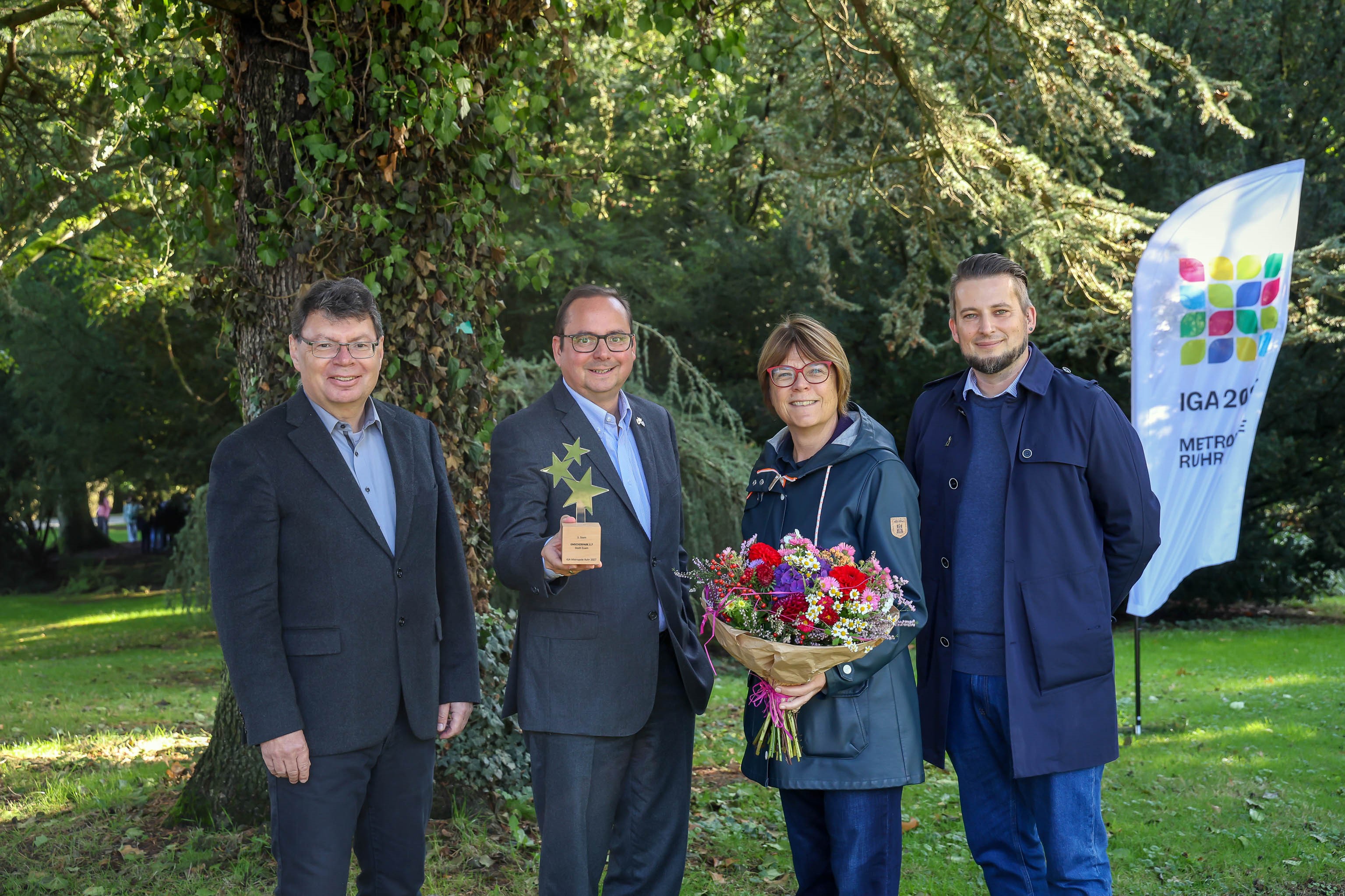 Horst Fischer (l.) überreicht Thomas Kufen, Simone Raskob und Mattis Ricken den dritten Stern und einen bunten Blumenstrauß der IGA 2027 für den Emscherpark 2.7. Im Hintergrund der IGA 2027 Aufsteller
