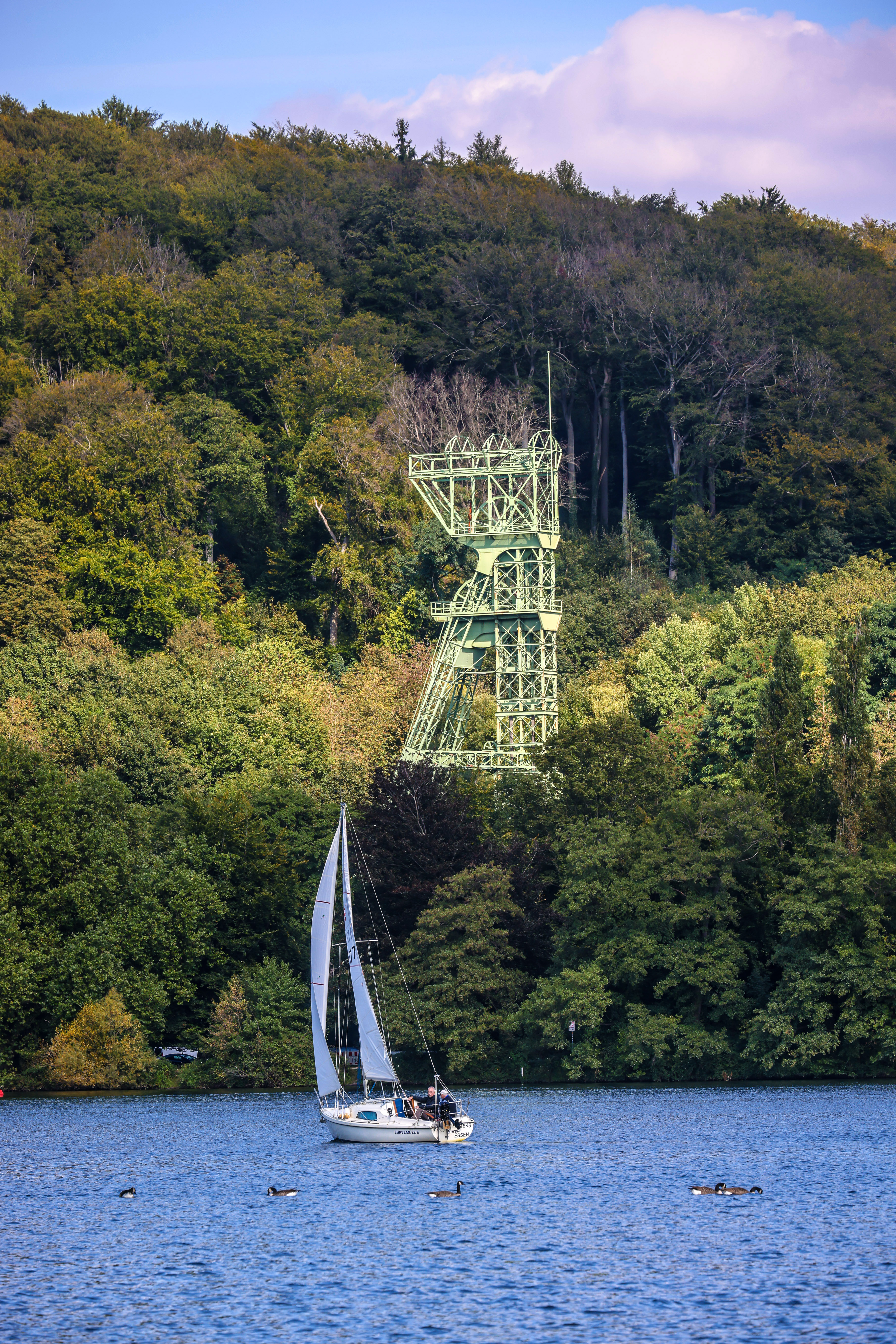 Im Vordergrund ein Segelboot auf dem See und dahinter ein grüner Förderturm bis zur Hälfte zugewachsen von Bäumen.