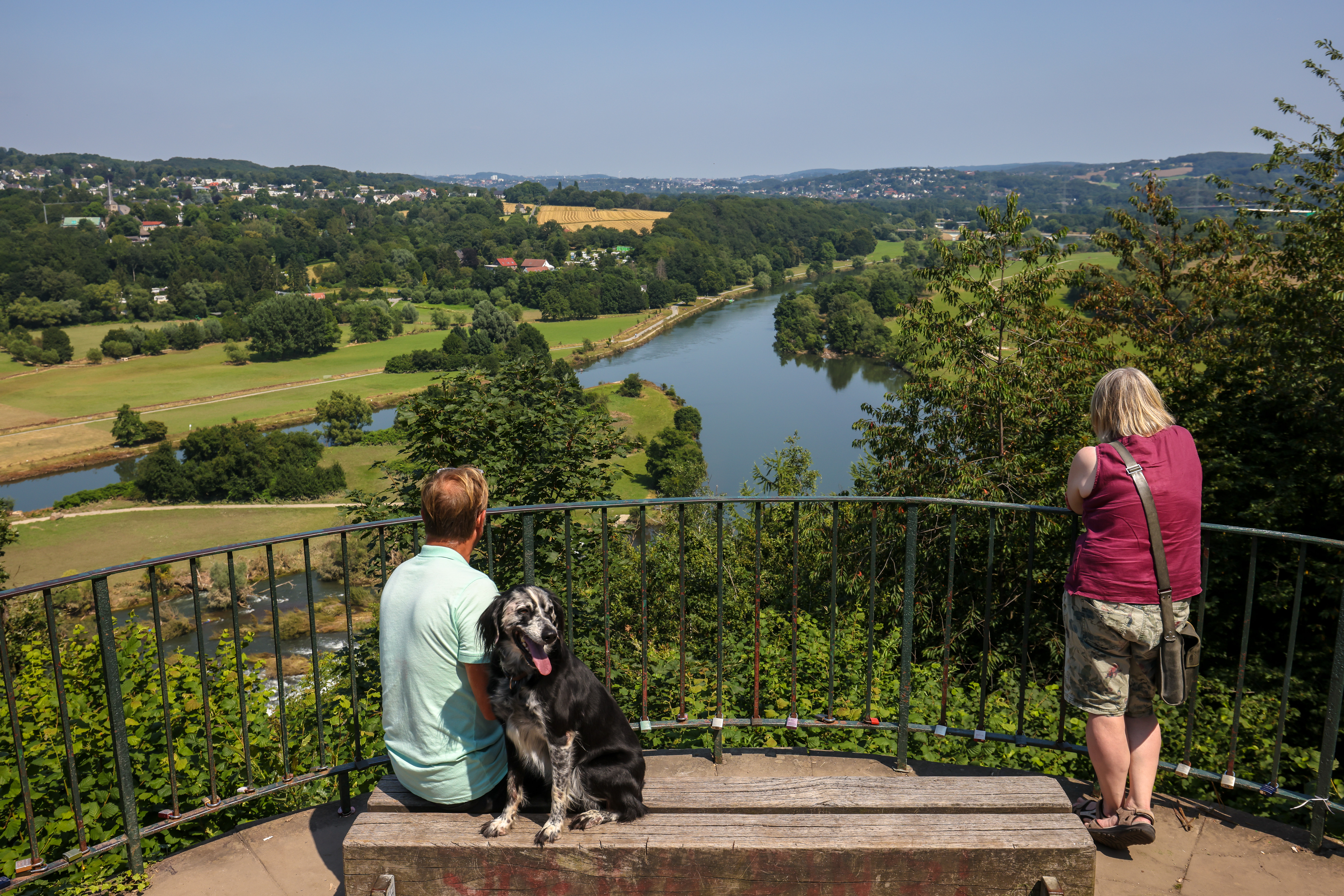 Ein Mann sitzt mit Hund auf einer Bank und eine Frau steht am Geländer. Beide blicken auf die Ruhr  umgeben von grüner Wiese, Bäumen und Häusern im Hintergrund.