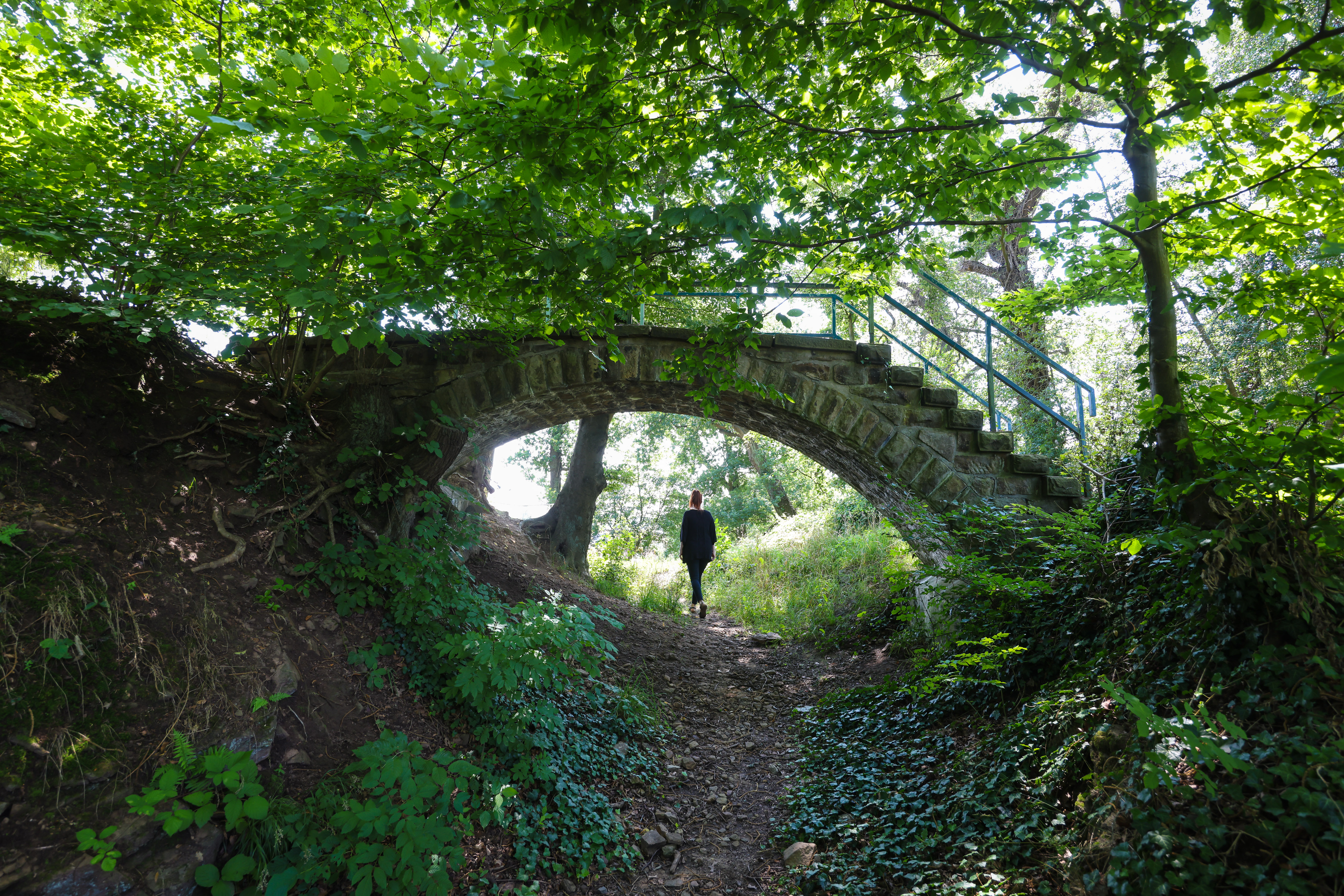 Eine Frau läuft unter einer Brücke aus altem Stein (mit Treppen) durch. Die kleine Brücke erstreckt sich über einen Waldweg und ist von Bäumen teils versteckt.