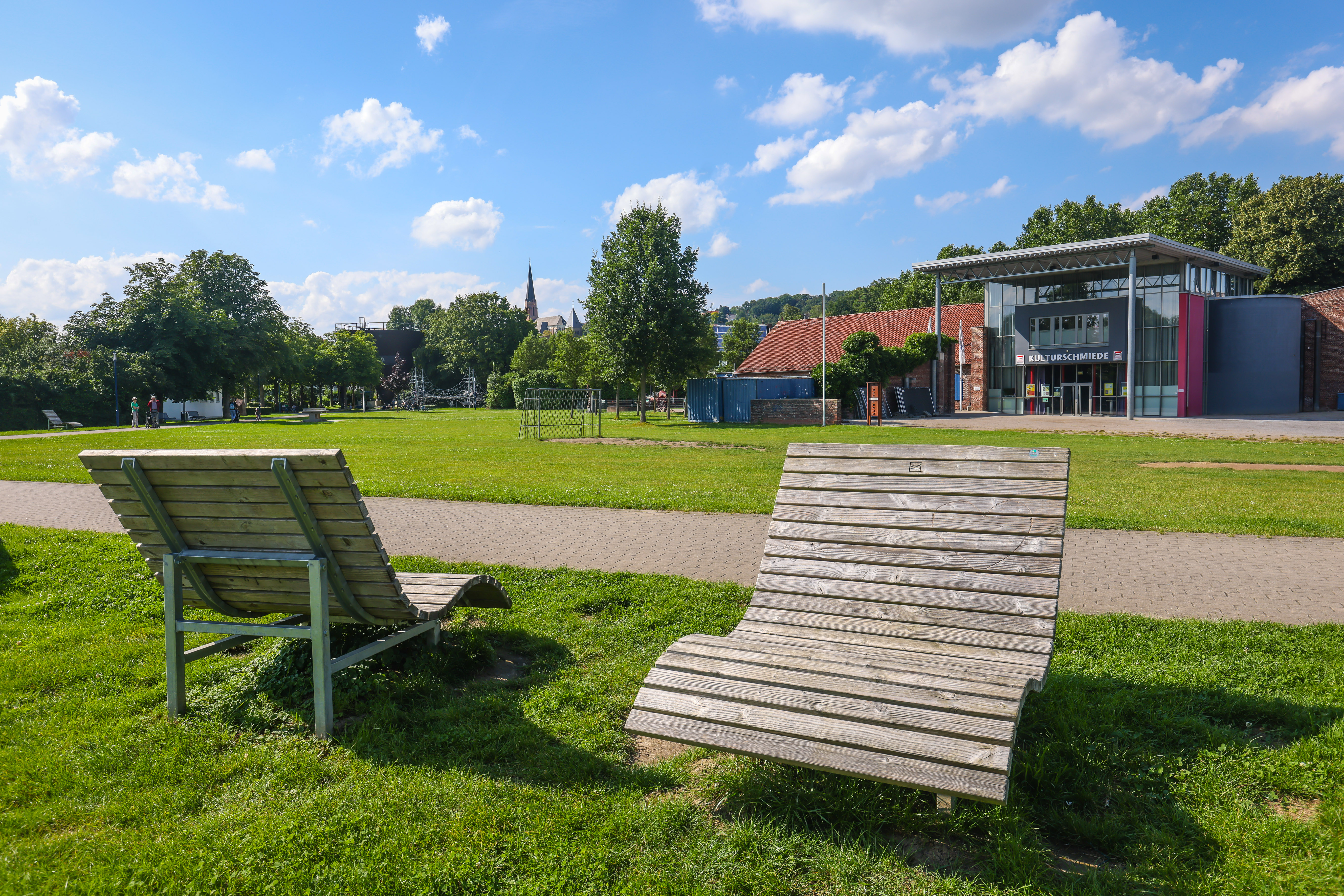 zwei Liegen aus Holz auf einer Wiese neben einem Fußgängerweg vor dem Kulturhaus