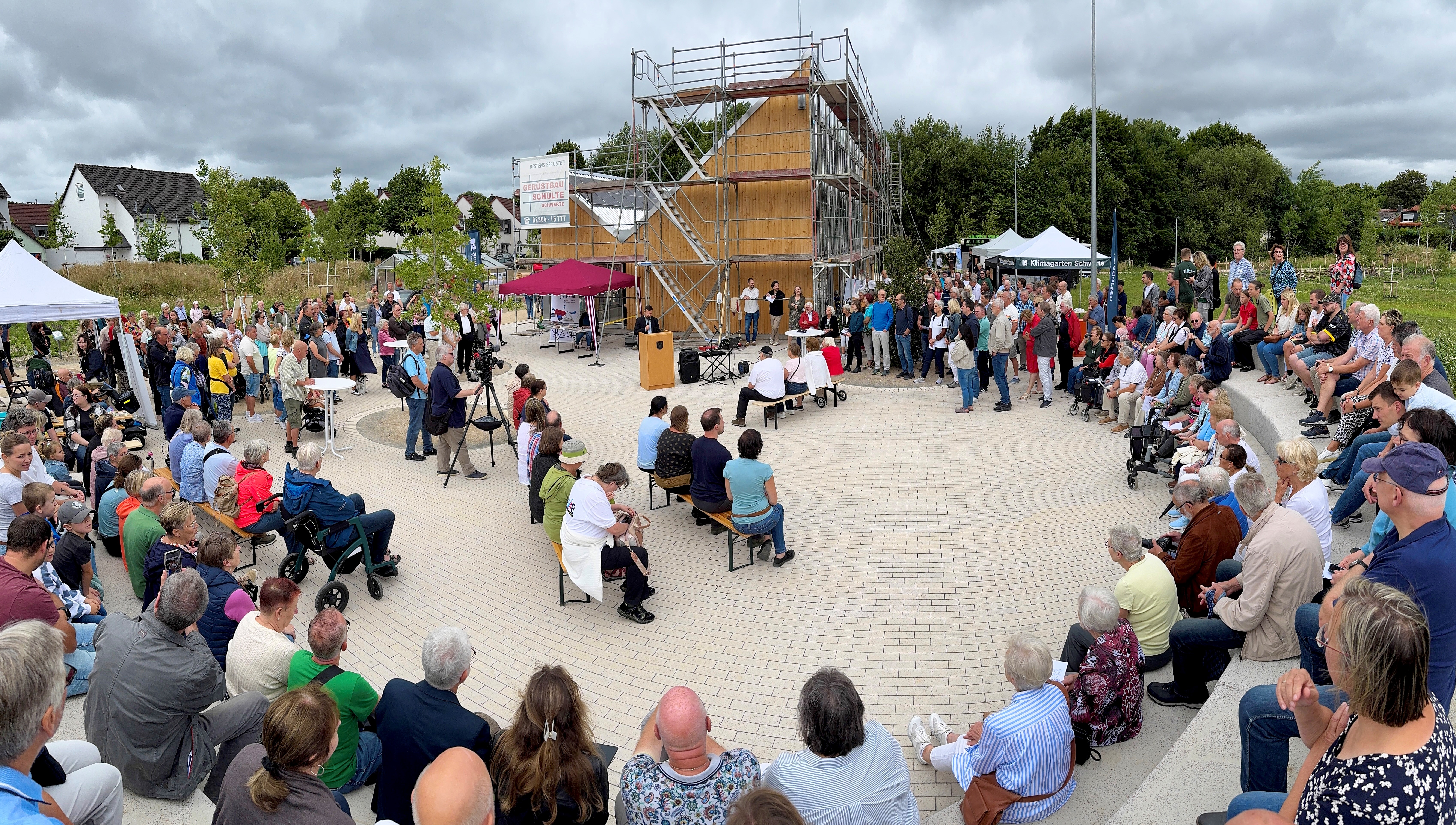 Viele Menschen sitzen und stehen im großen Kreis und nehmen an der Eröffnungsfeier für einen neuen Park teil. 