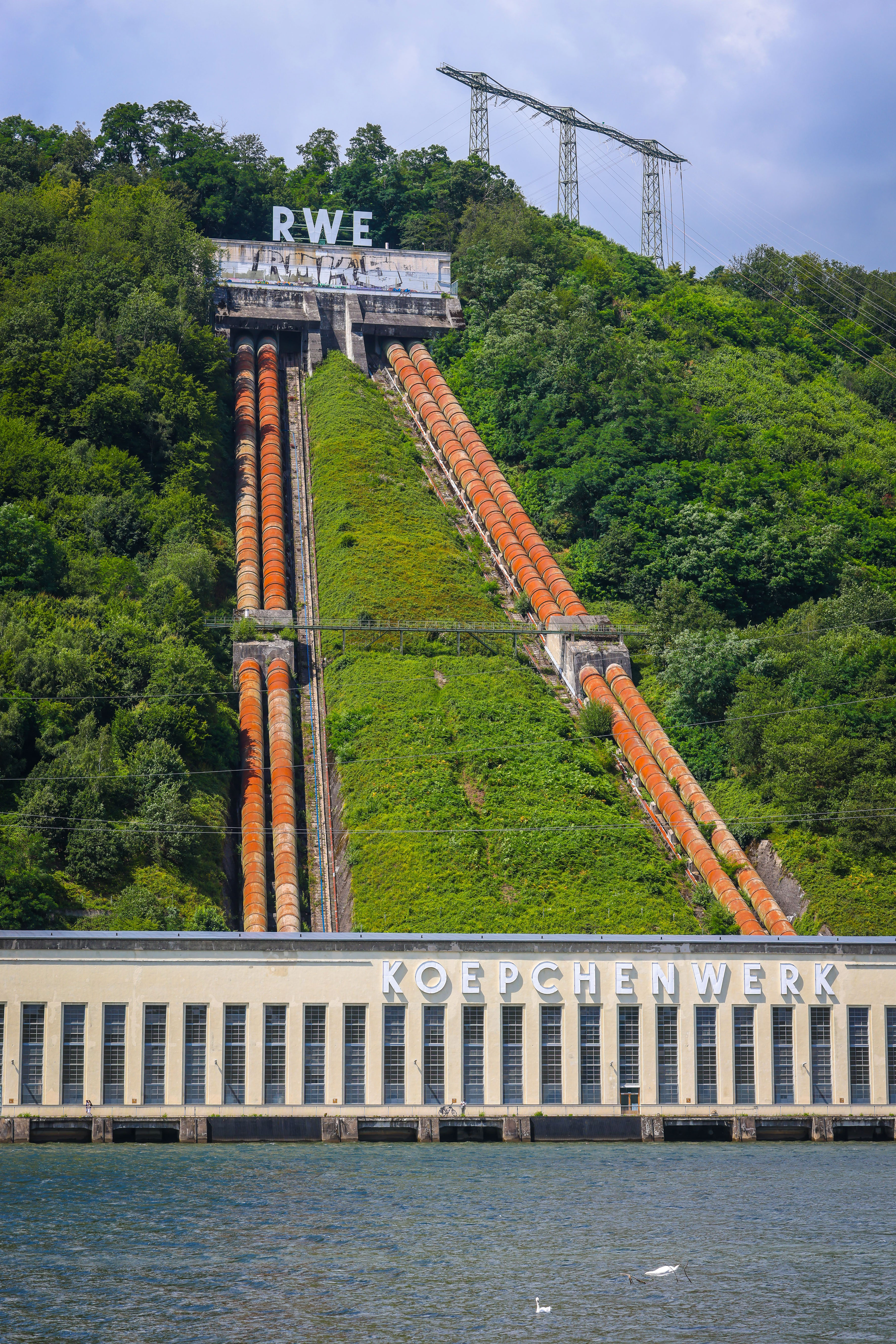 Blick auf das Koepchenwerk von der Ruhr aus. Vier große Rote Rohre führen vom unteren Werk den Hang hinauf.
