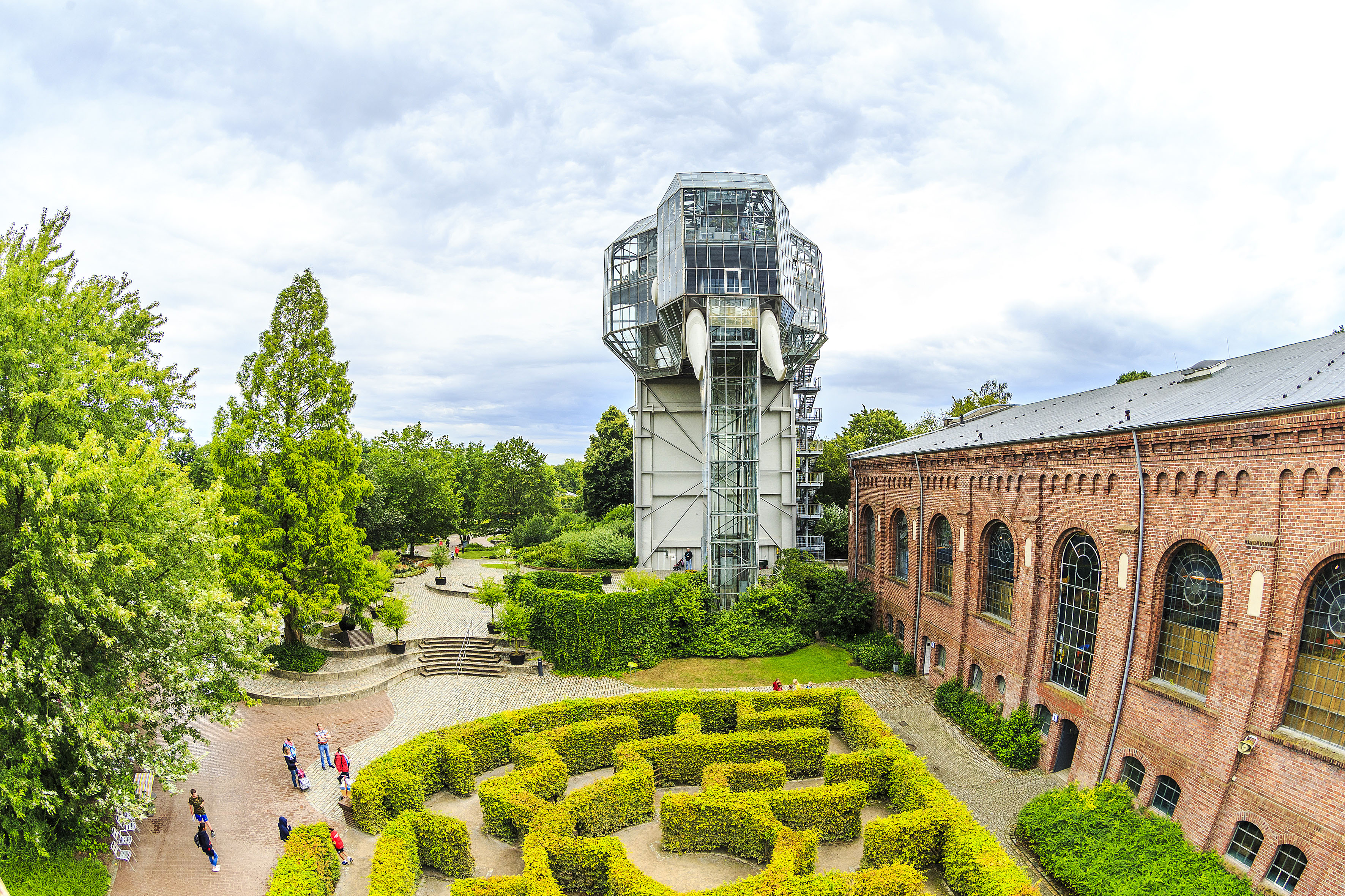 Ein Gartenlabyrinth zwischen hohen Büschen und rechts daneben ein Backsteingebäude