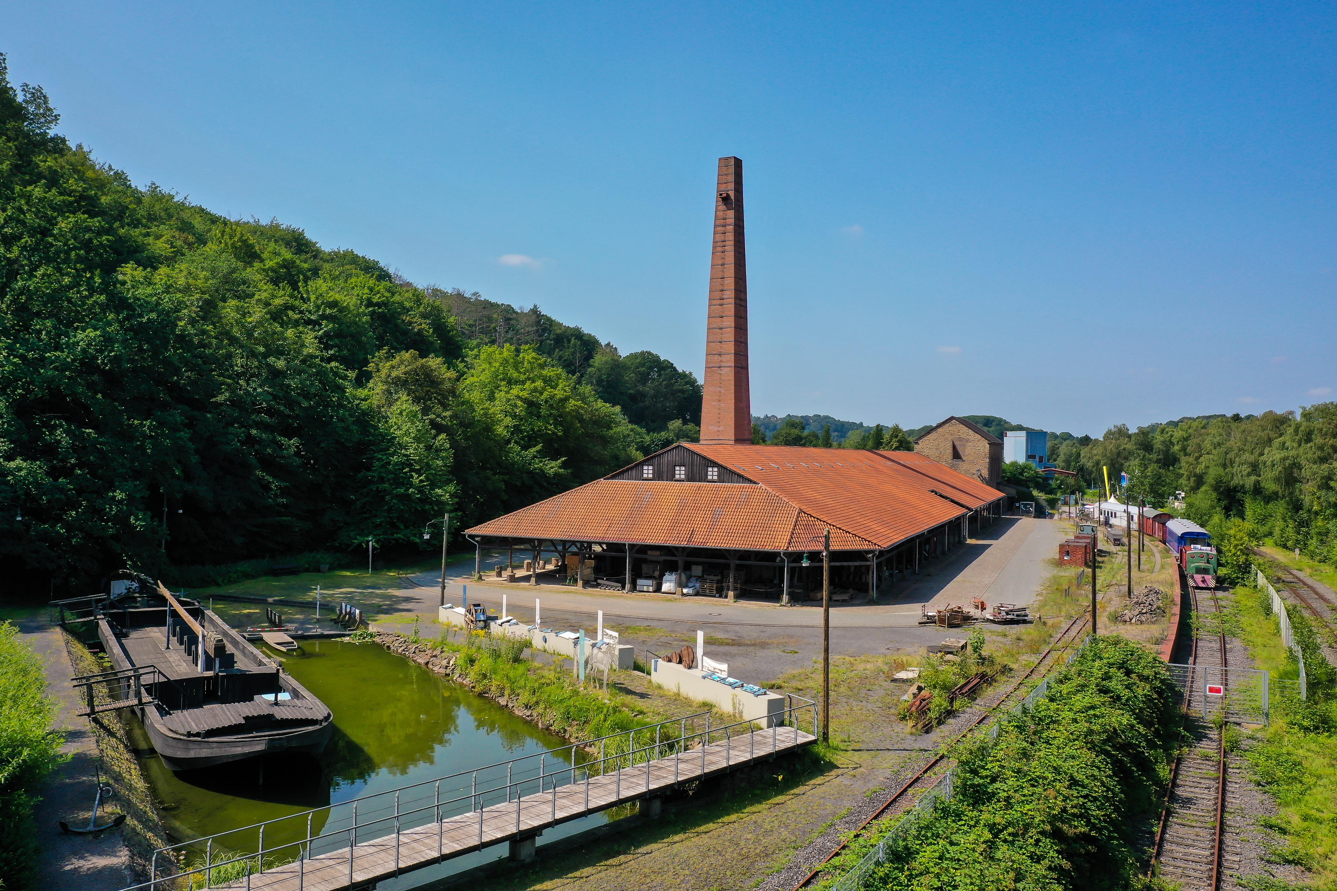 Blick auf die Zeche Nachtigall mit rot/braunem Schornstein, rechts Güterbahnhof, im Vordergrund ein Wasserbecken, links grüne Bäume