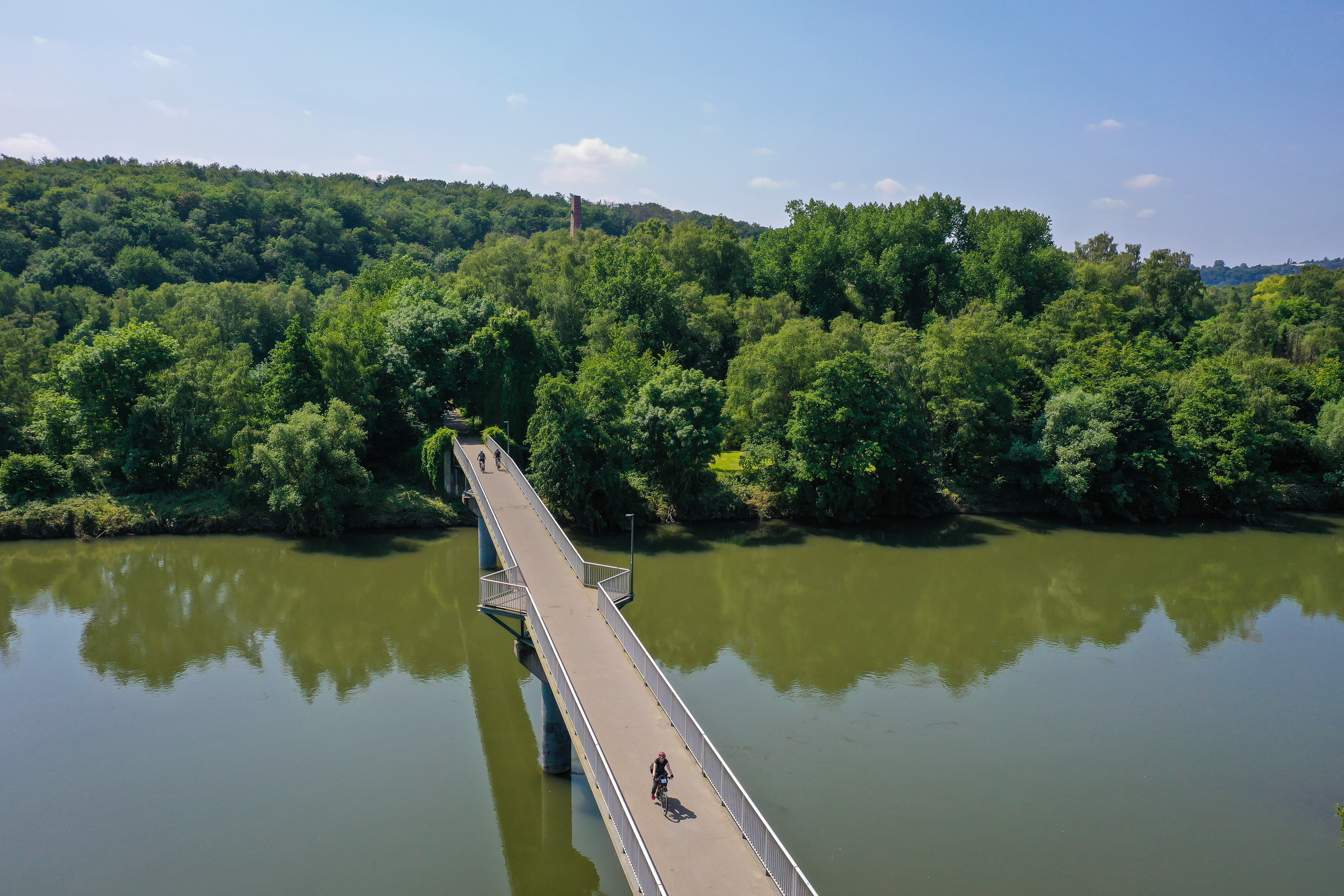 Eine Brücke mit Fahrradfahrern, die den Fluss überquert. Hinter dem Fluss dichte, grüne Bäume