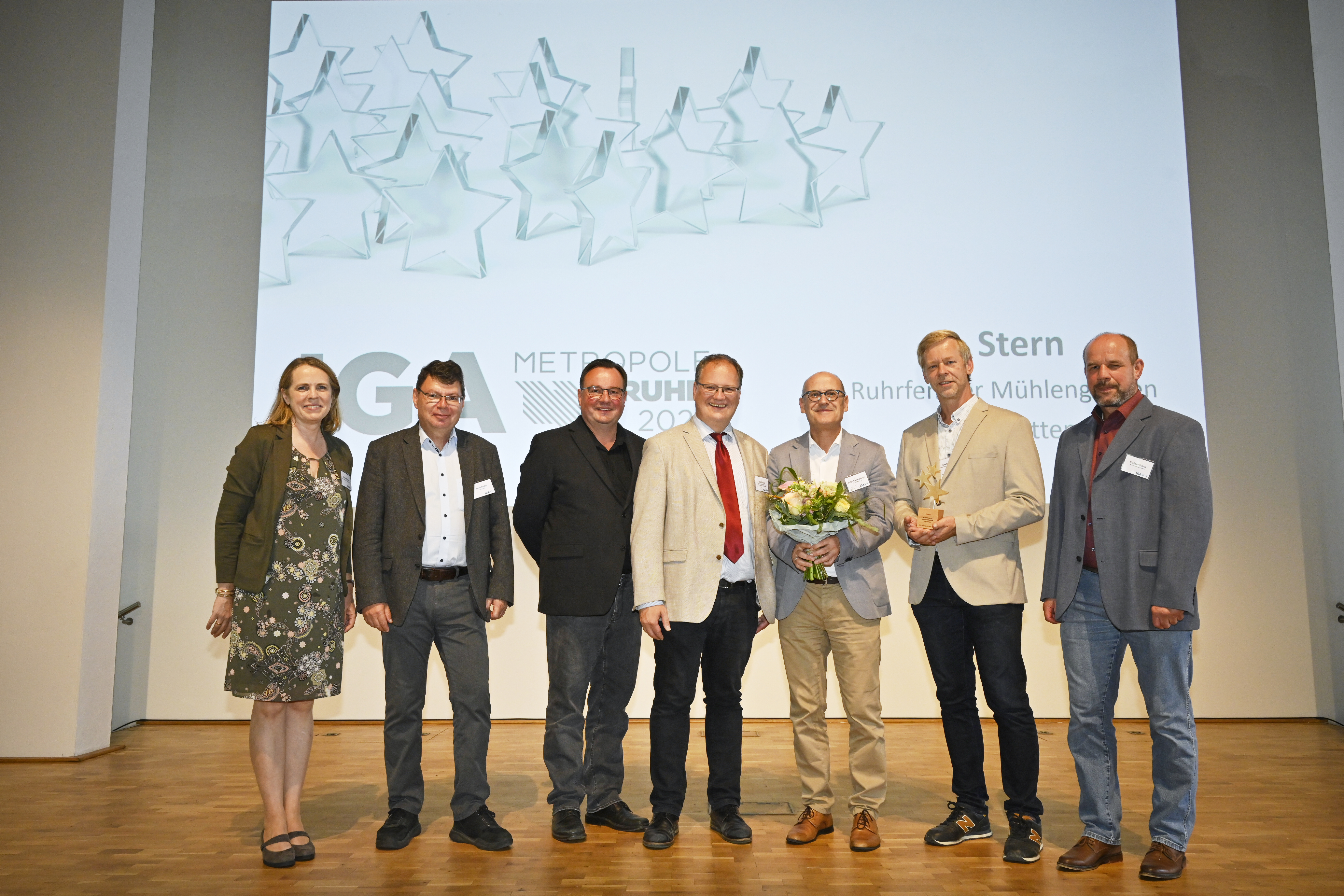Eine Frau und fünf Männer, davon einer mit feierlichem Blumenstrauß, vor der Leinwand "IGA Metropole Ruhr"