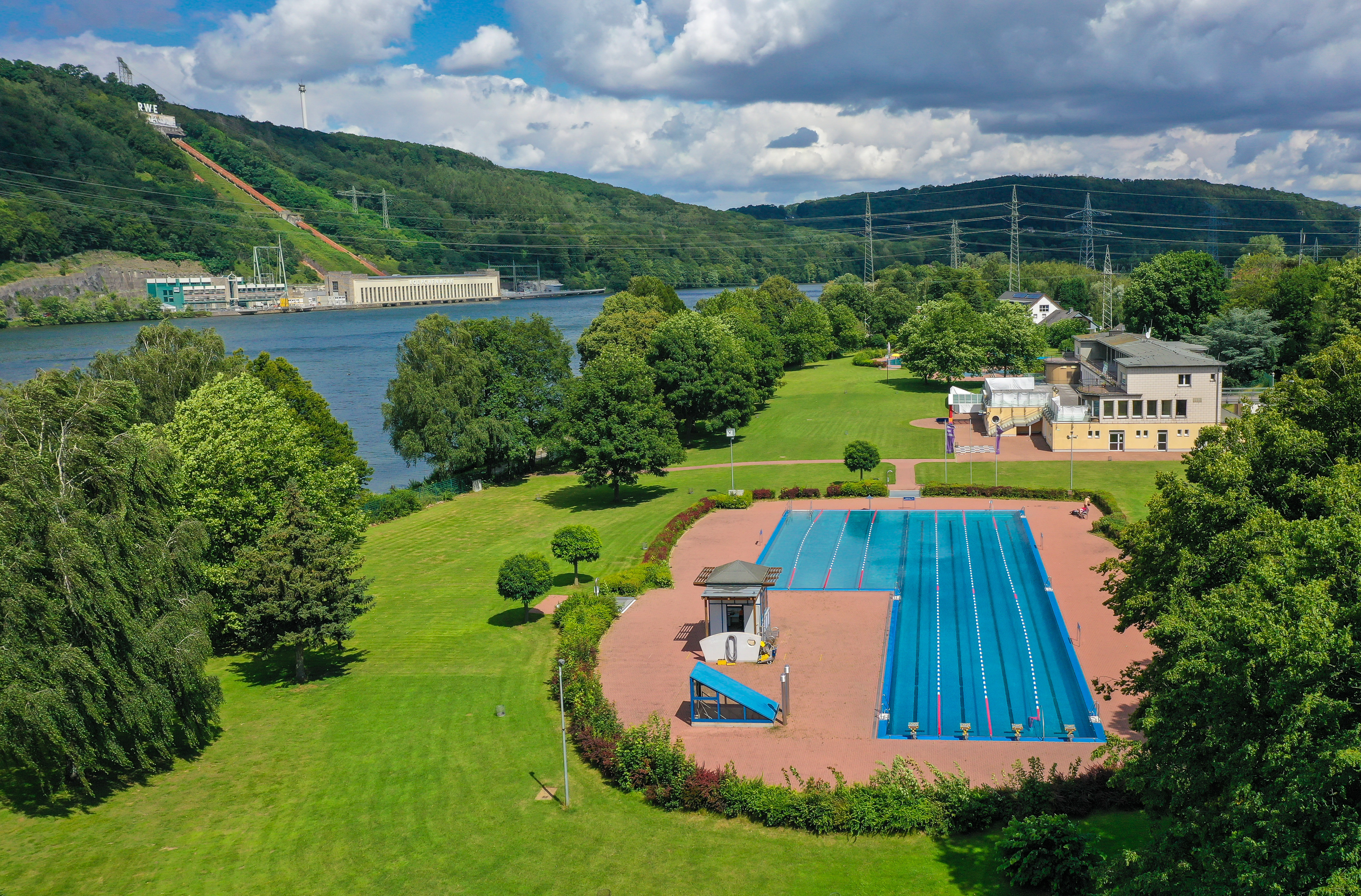 Vogelperspektive auf das Freibad umgeben von grünen Wiesen, direkt am Ufer des Hengsteysees. Im Hintergrund das Koepchenwerk auf der gegenüberliegenden Seite 