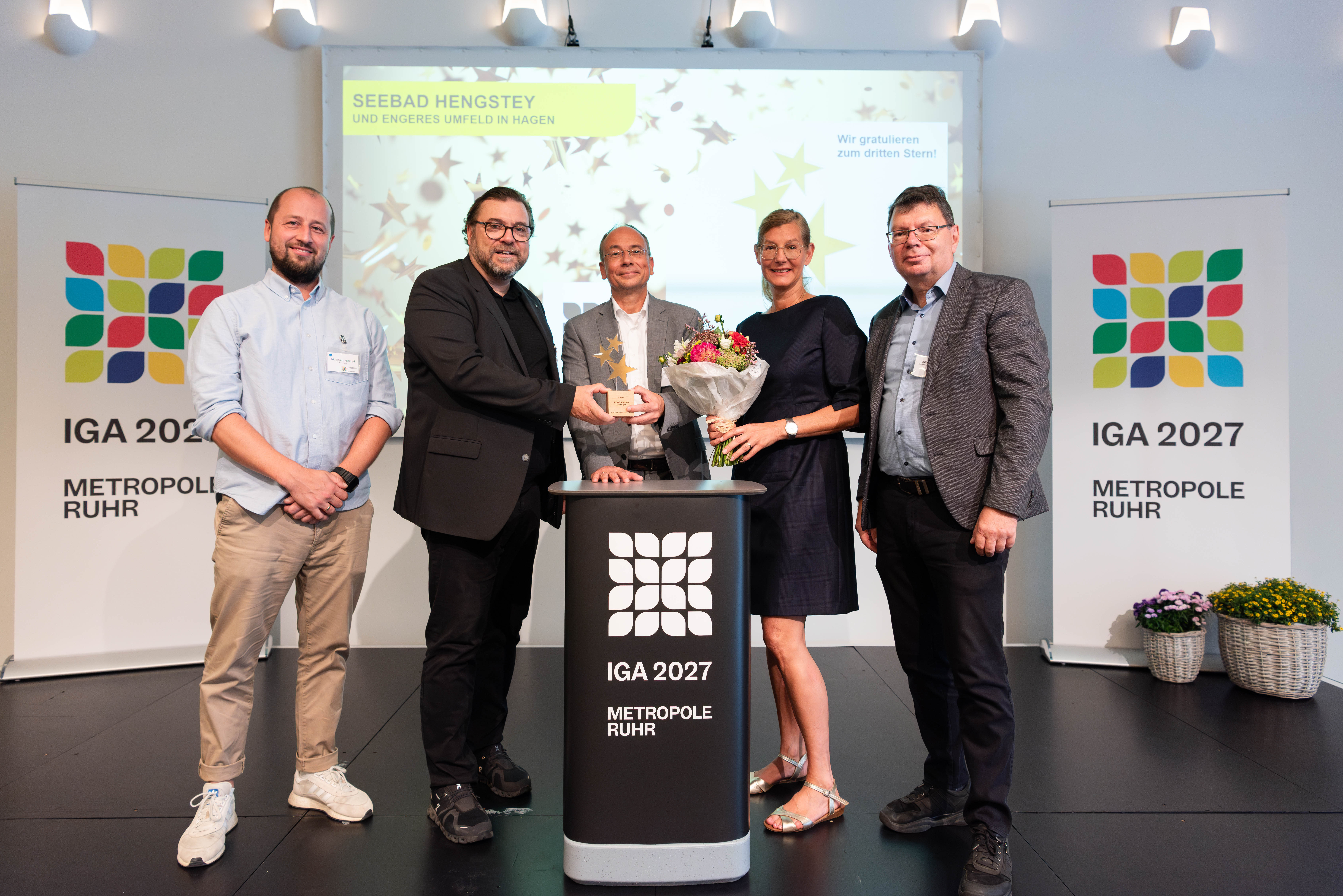 Fünf Personen vor IGA Plakaten bei der Überreichung der Trophäe und Blumenstrauß