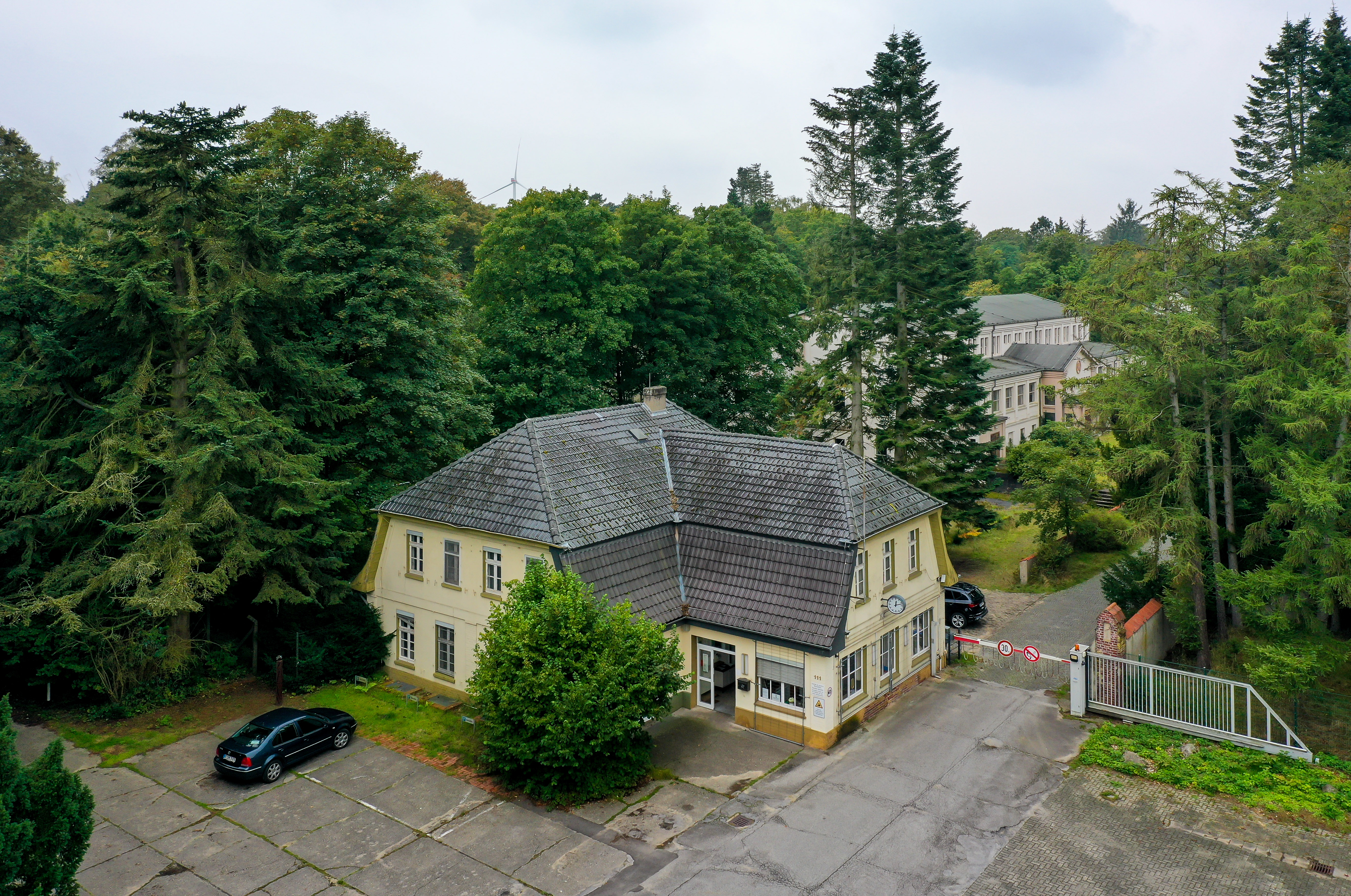 Blick aus Vogelperspektive auf das gelbliche Pförtnergebäude und Eingang mitten im Wald