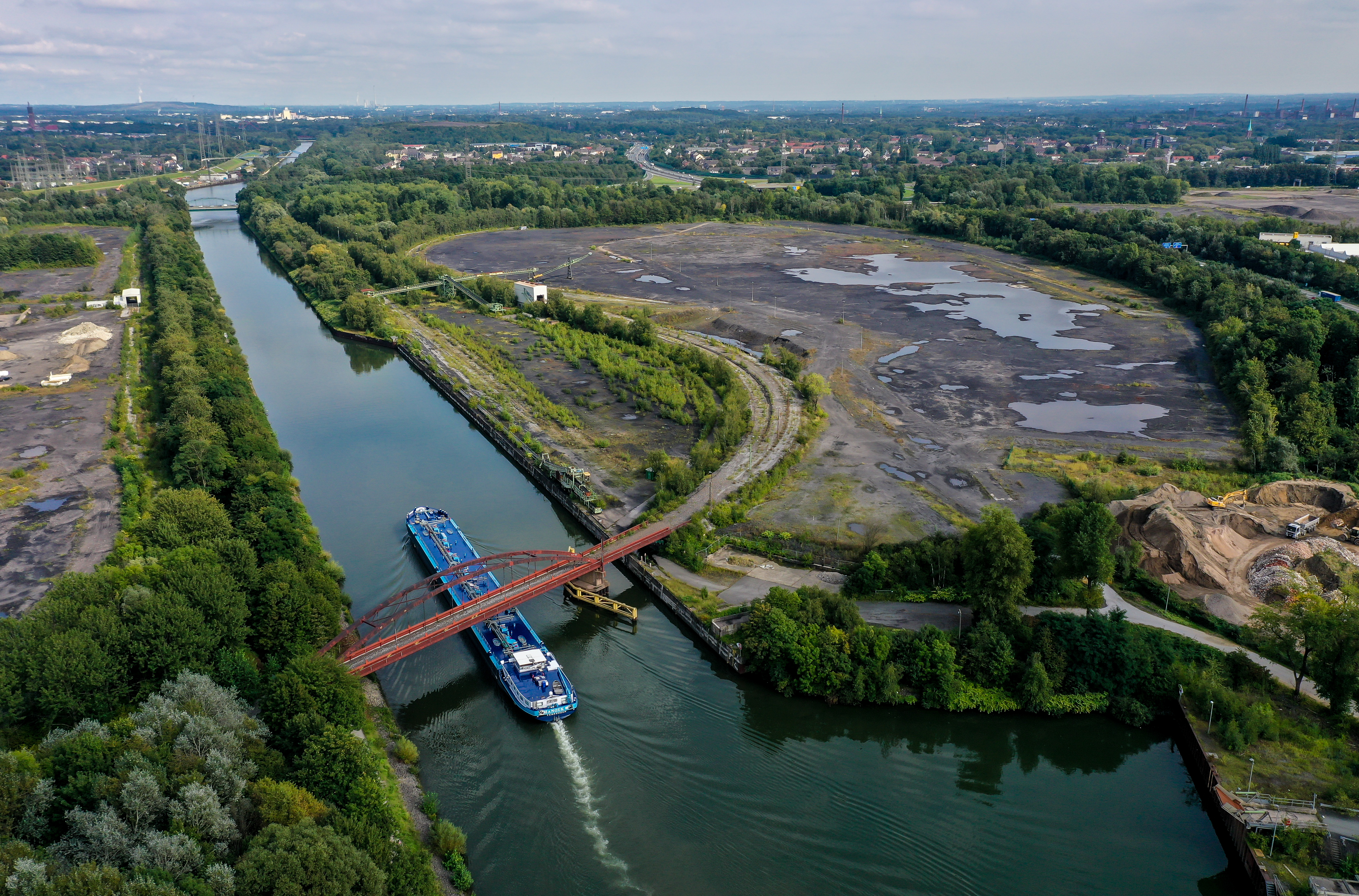 Vogelperspektive auf den Fluss mit roter Brücke und blauem Schiff. Links und Rechts neben dem Fluss große, ungenutzte Flächen.