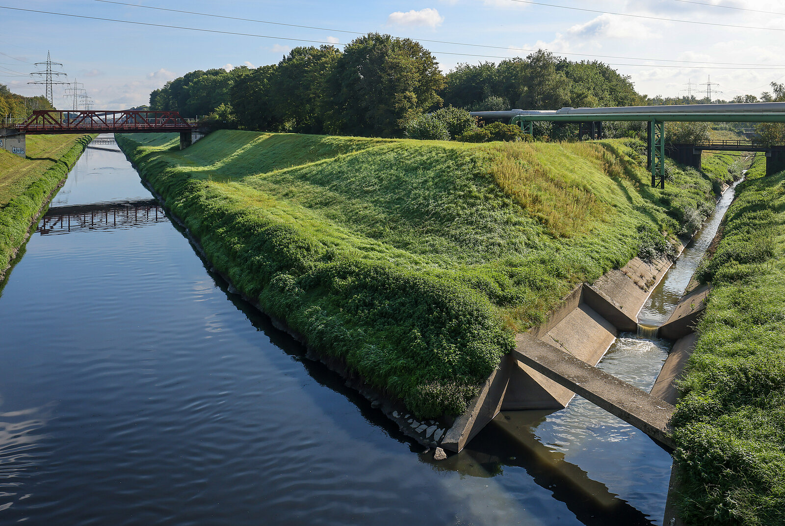 Der Fluss spaltet sich in zwei und rechts entsteht ein kleinerer, angelegter Bach; Links über dem Fluss ist eine rote Brücke. Umgeben ist der Fluss von grüner Wiese und Bäumen