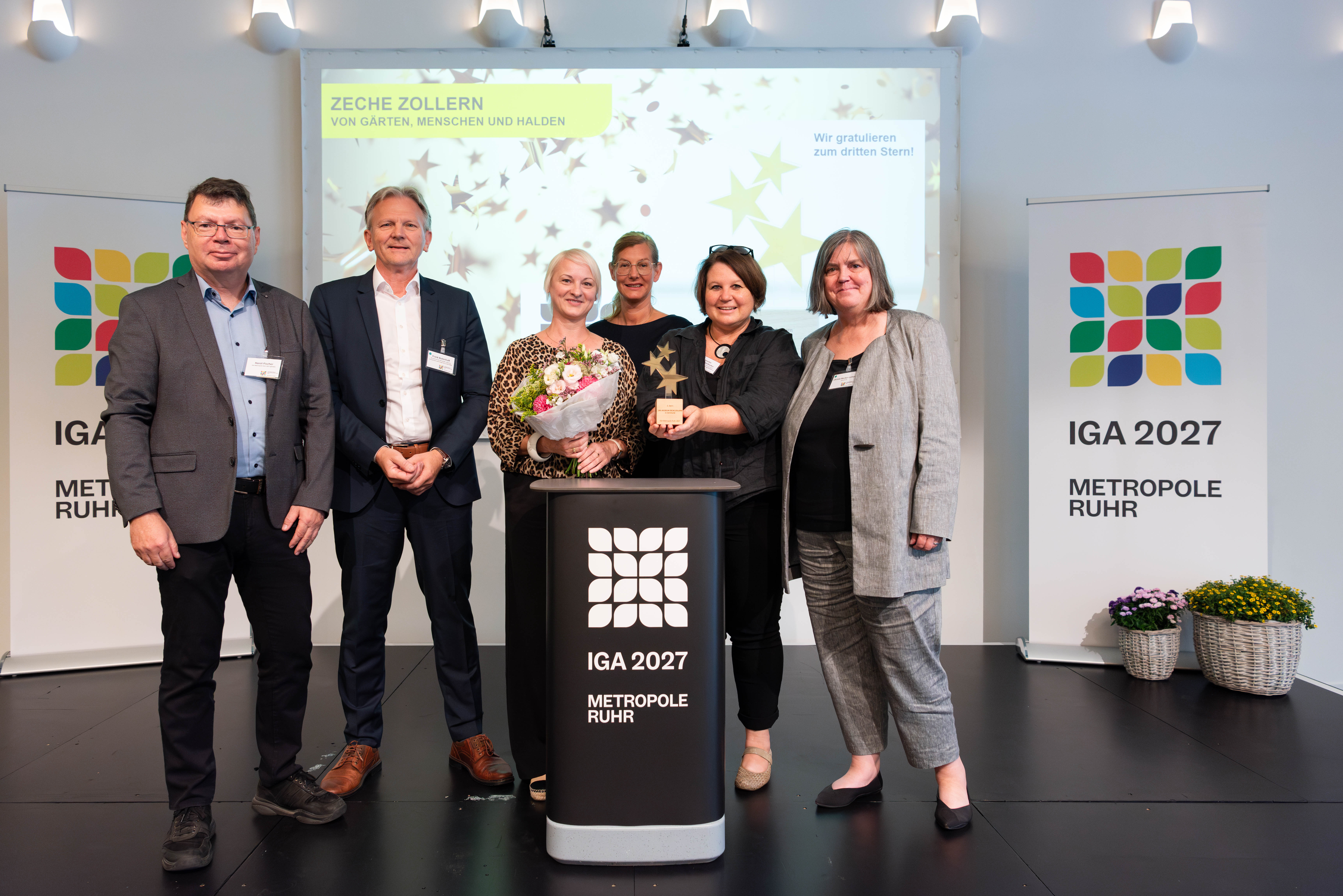 6 Personen mit Blumenstrauß und Sterne-Trophäe vor IGA 2027 Bannern